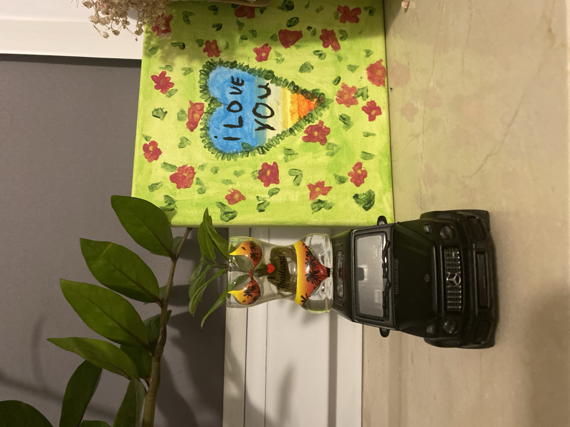 A black toy Mercedes G-Wagon is positioned on a beige marble countertop. Perched atop the toy car is a clear glass vase shaped like a female torso wearing a bikini, containing a small green plant with multiple leaves. To the right of the plant vase, a square canvas is leaning against a wall. The canvas is painted lime green with a repeating pattern of red flowers and green leaves. In the center of the canvas, a heart shape is drawn with a blue interior containing the text "I LOVE YOU" in black lettering. The bottom of the heart features a gradient of orange and yellow. To the upper left of the canvas, a branch with several green leaves extends into the frame from the left side. The background wall is a solid gray. The scene is lit from the front, casting shadows behind the objects. No specific location in Łódź, Poland is discernible from the image.