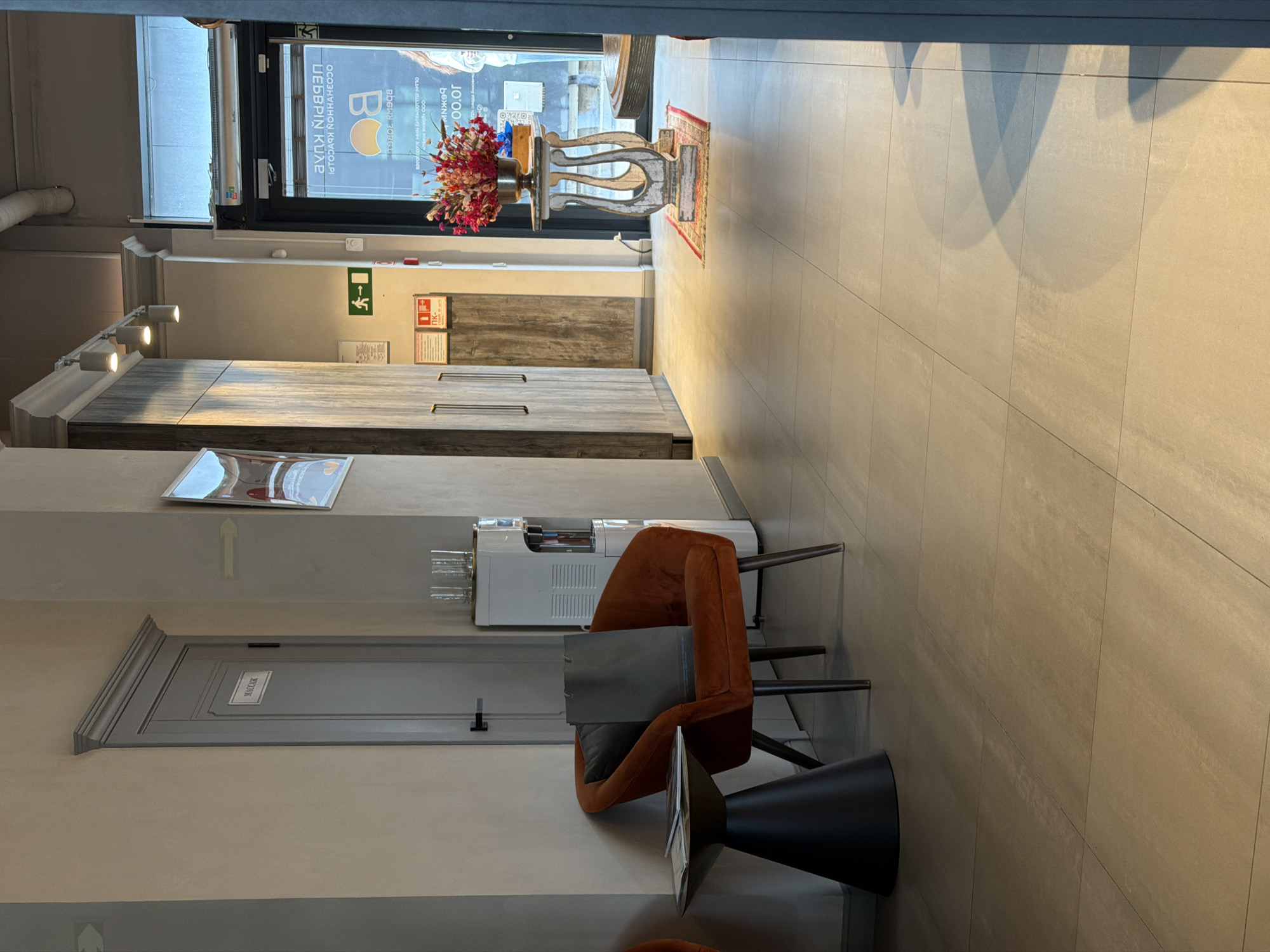 The image depicts a waiting area in a commercial establishment, possibly a beauty salon or spa, in Moscow, Russia. A modern, plush orange armchair is positioned to the left, with a dark gray bag and a small table with magazines beside it. Adjacent to the armchair is a water cooler. Further down the corridor, a door with a sign reading "МАССАЖ" (Massage) is visible. The environment is clean and well-lit, with tiled flooring and muted wall colors. A large glass door at the end of the hallway reveals a street scene and signage in Russian, indicating the establishment's location. A decorative vase filled with flowers stands on a pedestal near the entrance. The overall impression is of a professional and welcoming space.