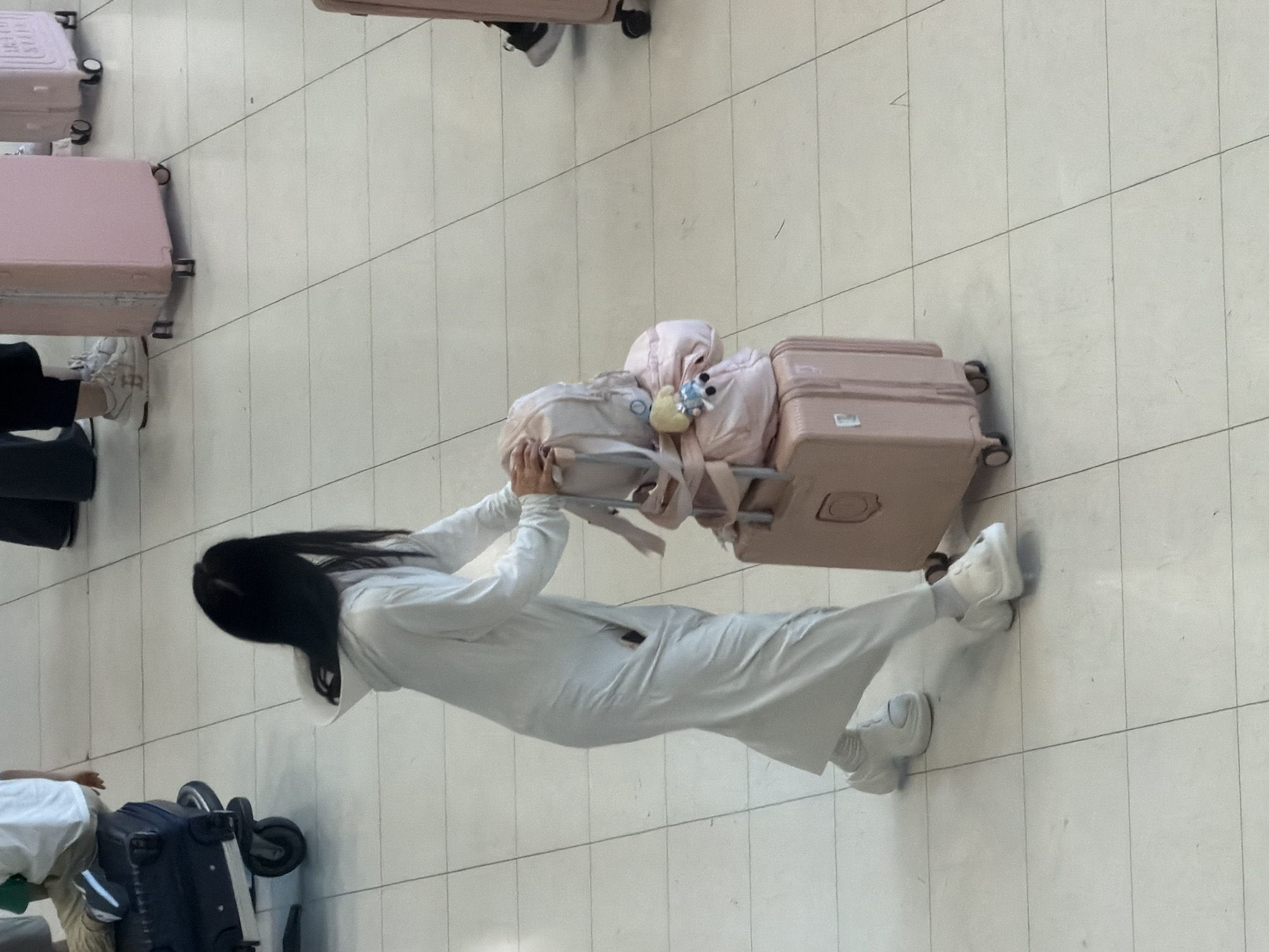 The image depicts a person in a white hooded robe and white sneakers, pulling a pink suitcase. Stacked on top of the suitcase are two pink bags, one of which has a small blue and blonde doll attached to it. The person is walking on a tiled floor, likely within an airport or a large public indoor space, given the presence of other luggage in the background, including a dark blue suitcase on a trolley to the left. The lighting suggests an indoor environment. The location is specified as Nong Prue Subdistrict, Thailand.