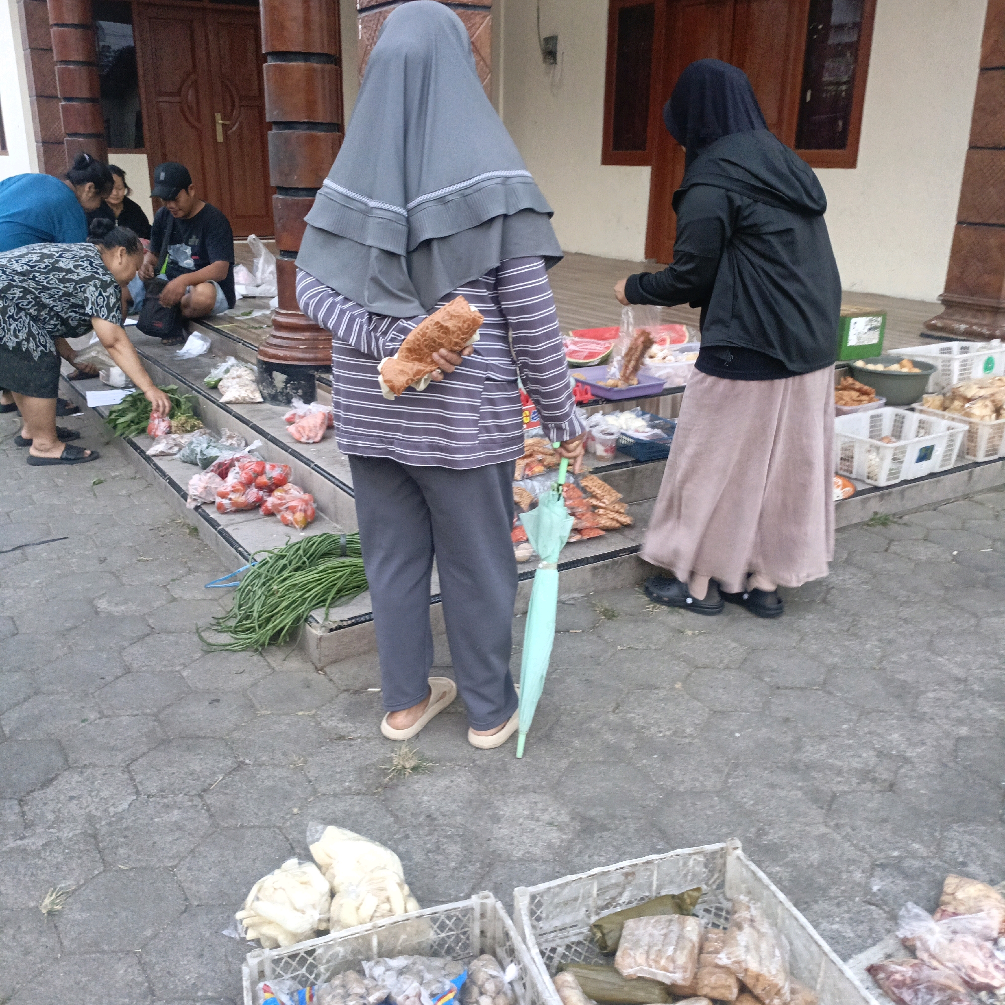 In the city of Salatiga, Indonesia, a group of people are gathered in front of a building entrance. The scene appears to be an outdoor market or roadside stall. Two women are visible in the foreground, one wearing a grey hijab and a striped long-sleeved shirt, holding a piece of food. The other woman, also in a hijab, is wearing a black jacket and a long skirt. They are positioned near crates filled with various food items. In the background, several other individuals are seated on steps leading up to the building. One man, wearing a black t-shirt and a cap, is interacting with a woman. The ground is paved with hexagonal stones. Crates containing produce like tomatoes, long beans, and other packaged food items are arranged on the steps and the ground. A light green umbrella is leaning against the leg of the woman in the striped shirt. The architectural style of the building suggests it might be a traditional Indonesian house or shop.