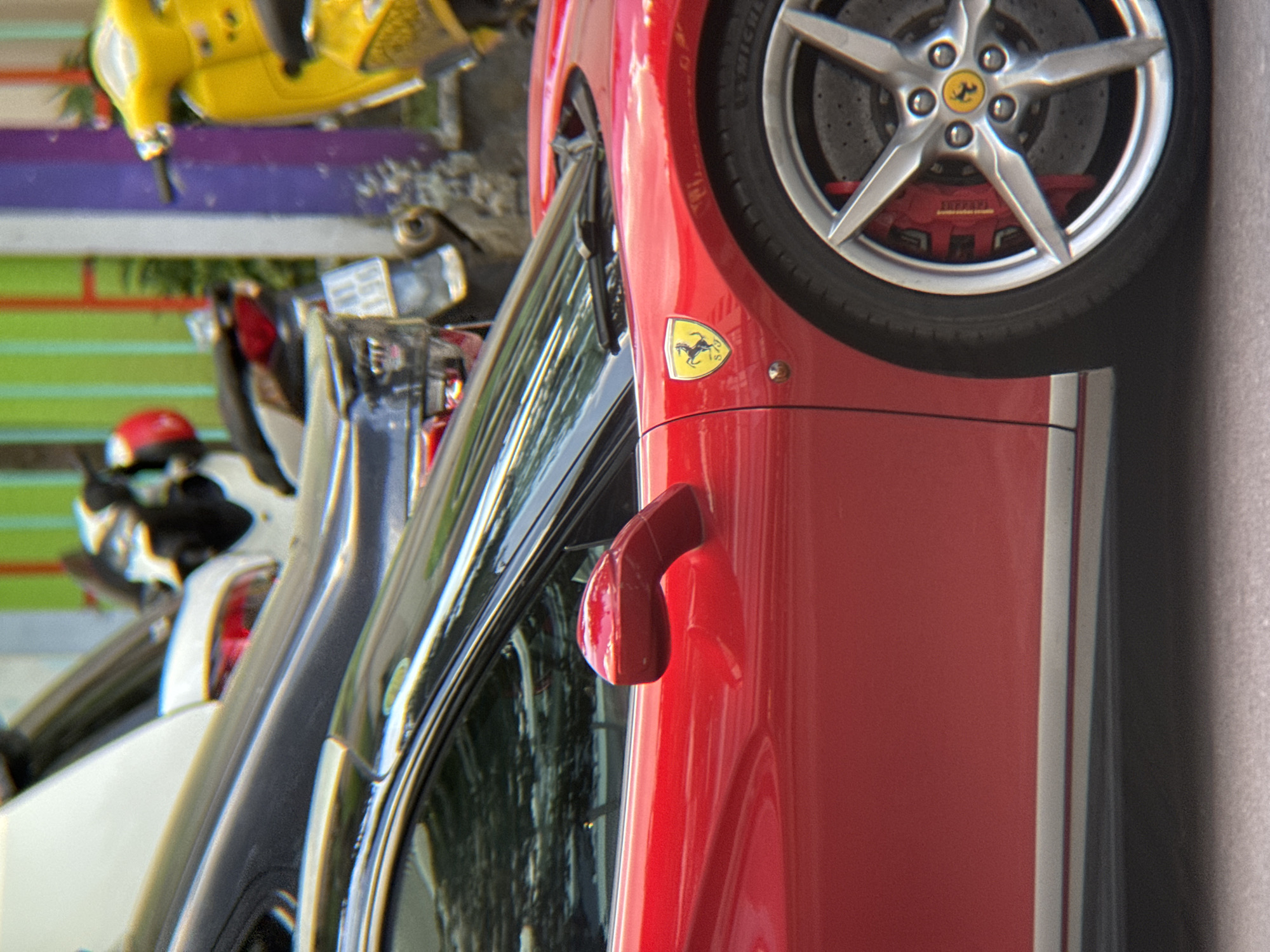 The image shows a close-up of a red Ferrari parked on a street in Nha Trang, Vietnam. The car is the primary subject, with its distinctive prancing horse emblem visible on the fender. The background features a colorful wall with vertical stripes of green, yellow, and purple, suggesting a vibrant urban setting. A yellow scooter is parked to the right of the Ferrari, and parts of another dark-colored vehicle are visible behind it. The street surface appears to be asphalt. The lighting suggests it is daytime, with bright sunlight reflecting off the car's polished surface.