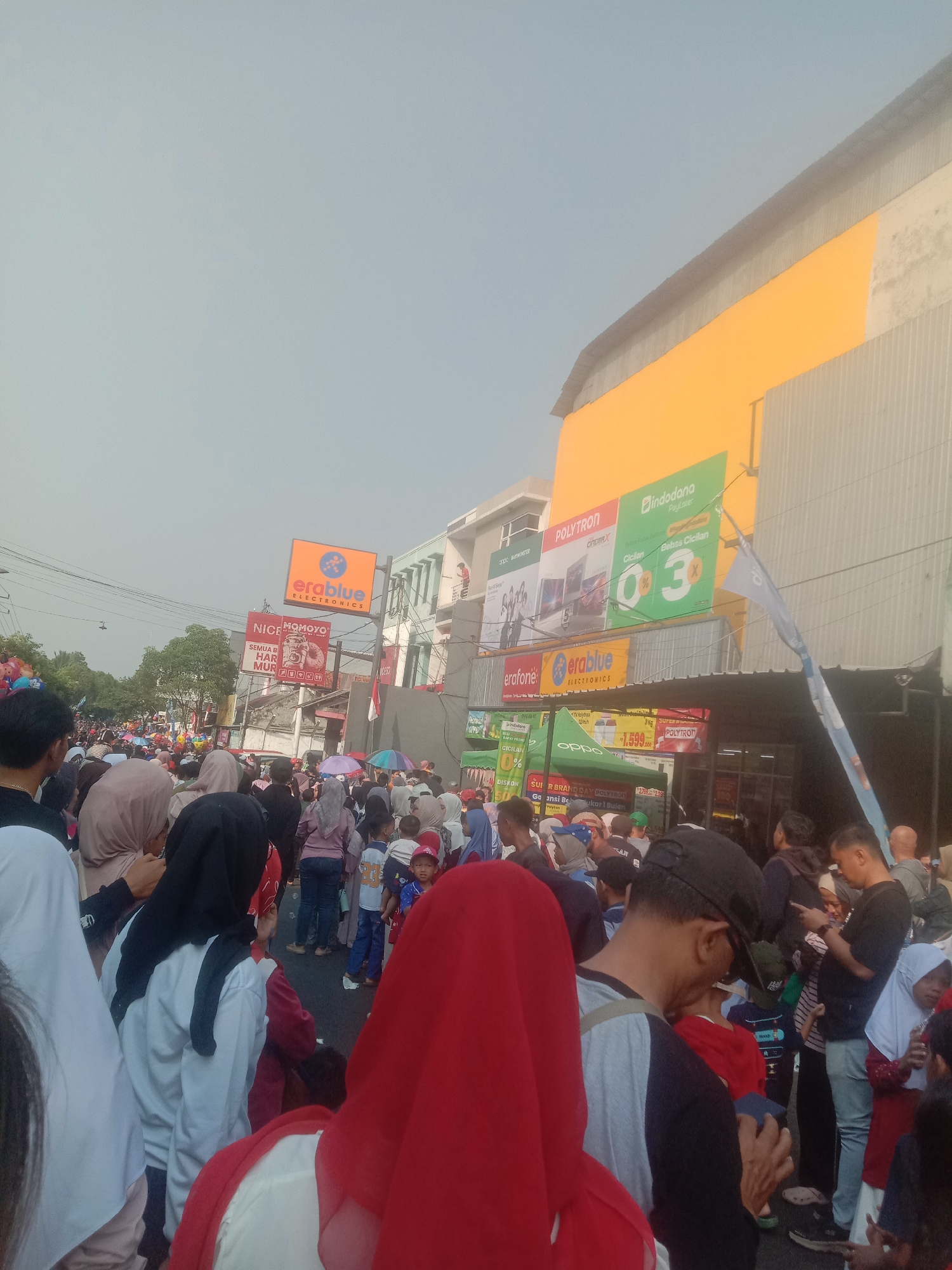 The image depicts a crowded street scene in Cianjur, Indonesia, likely during a daytime event given the bright sunlight. A large group of people are gathered, many wearing casual clothing, with a significant number of women and children present. Some individuals are wearing headscarves. The background features several multi-story buildings with prominent signage. One building displays large advertisements for electronics brands like "POLYTRON," "erablue ELECTRONICS," and "Indodana PayLater," indicating the presence of retail establishments. Another sign reads "NICE MOMOYO." Tents are set up along the street, suggesting a market or promotional event. A blue flag is visible on the right side of the image. The overall atmosphere appears to be one of public assembly and commerce.