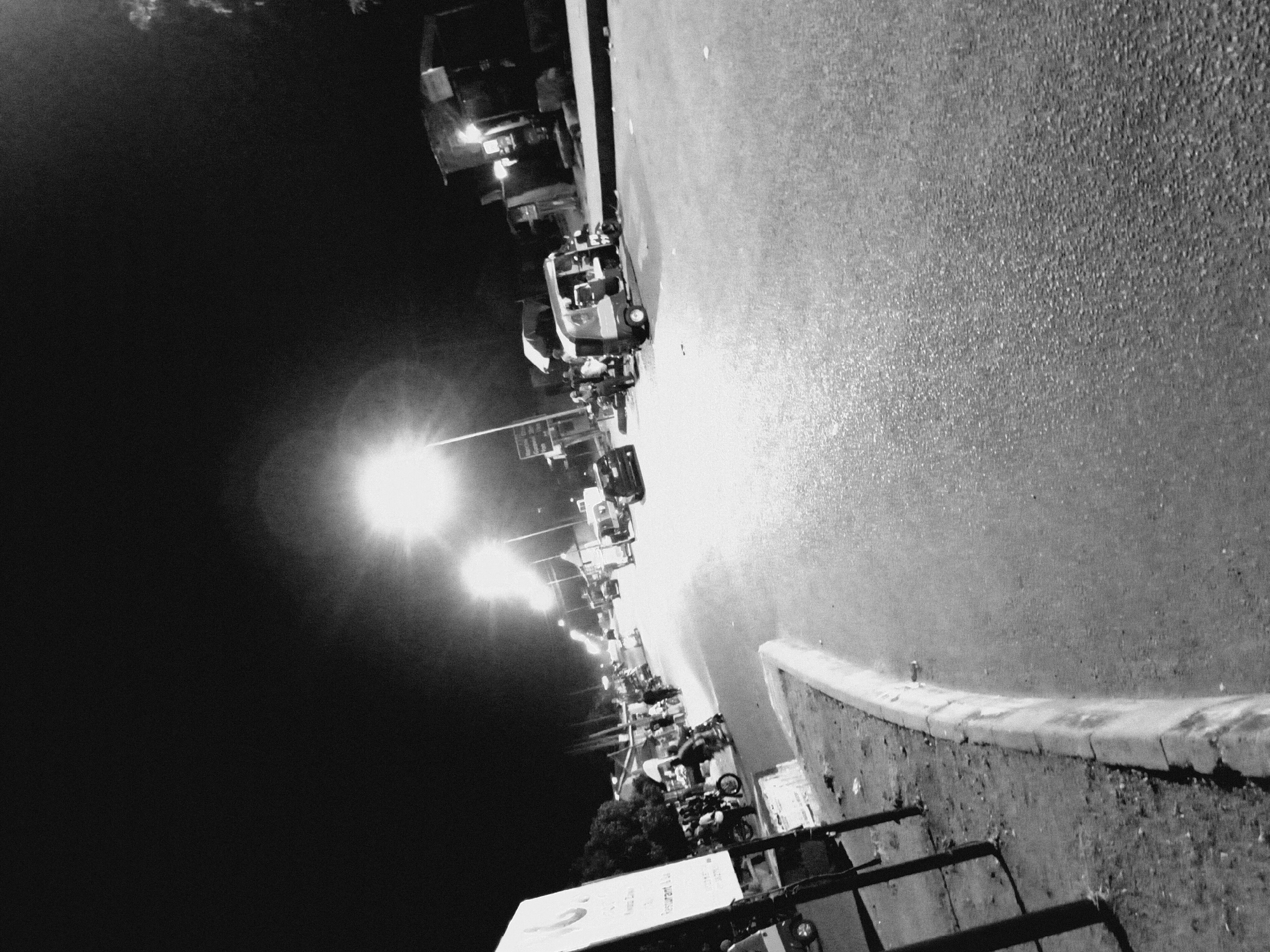 The image is a black and white, low-light photograph taken at night on a street in Bukuru, Nigeria. The scene depicts a busy street with several vehicles, including a few auto-rickshaws (also known as 'keke napep'), motorcycles, and at least one car. Pedestrians are visible in small groups, some near the vehicles, suggesting a public transportation hub or a marketplace. Streetlights illuminate the scene, casting long shadows and creating a slightly grainy texture due to the low light conditions. On the left side of the image, a signpost with text is partially visible, along with some foliage and a curb. The road surface appears to be asphalt and shows some wear and tear. The overall impression is of a lively, albeit dimly lit, urban environment at night.
