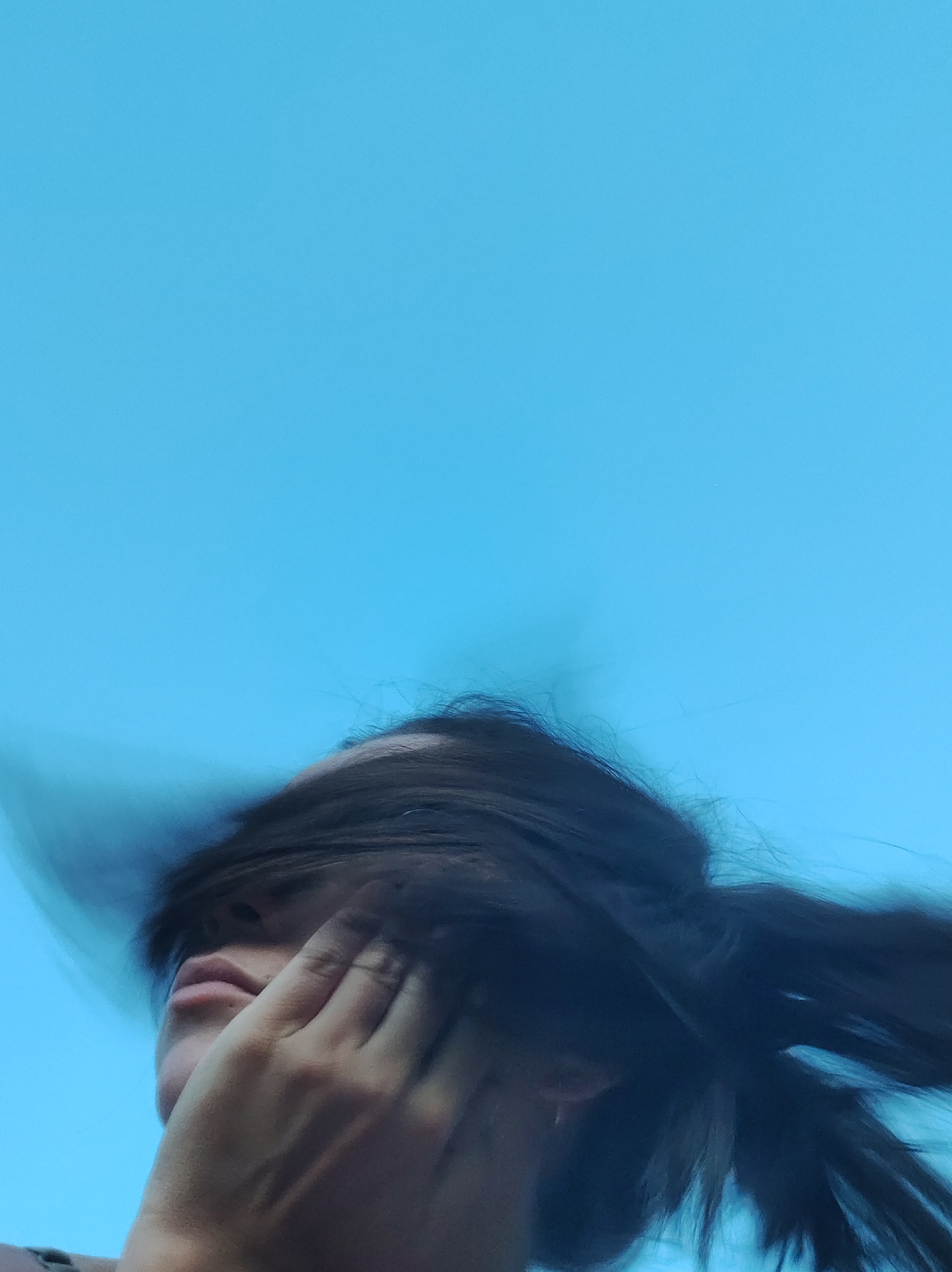 A low-angle, upward shot captures a person's face partially obscured by their dark, windblown hair, against a clear, bright blue sky. The person's right hand is raised, with fingers pressed against their face, covering their eye and part of their forehead. Their lips are slightly parted, and their nose is visible. The background is a solid expanse of sky with no discernible landmarks or features. The image is set in the city of Фоки, Russia.