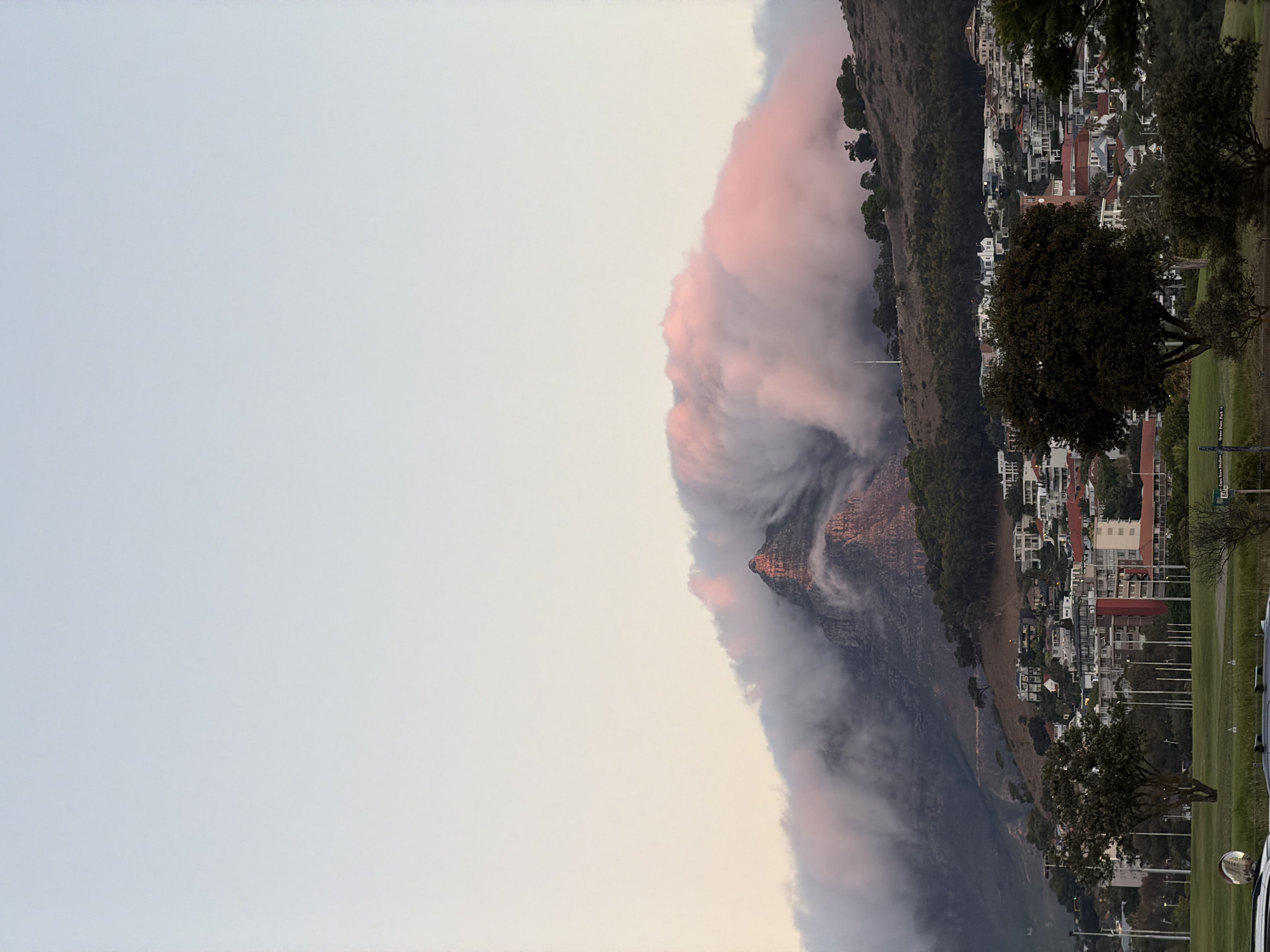 The image shows Lion's Head mountain in Cape Town, South Africa. A thick blanket of clouds is draped over the mountain. In the background, there are buildings and trees. The sky is a pale grey.