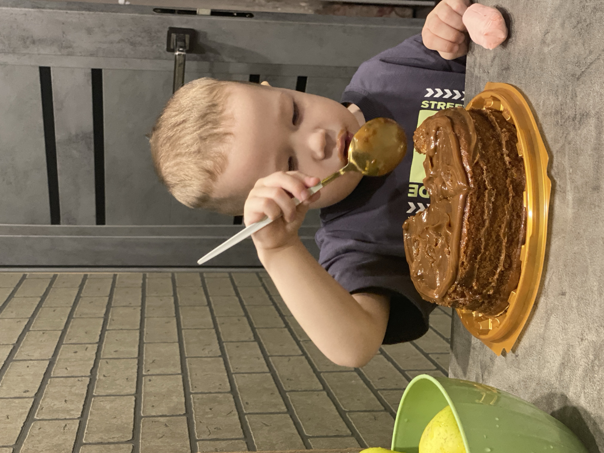 A young child with light hair is captured in a close-up, rotated image, likely indoors in Sochi, Russia. The child, wearing a dark blue t-shirt with partial white text "STREE" and "DE", is actively eating a chocolate cake with a golden spoon. The spoon is held in his right hand and has a white handle, currently filled with chocolate frosting. Some chocolate is visible around his mouth.
The cake is a rich dark brown, heavily frosted, and sits on a golden plastic tray on a dark gray, textured countertop or table surface. To the lower right, a green bowl containing yellow fruit, possibly lemons or apples, is partially visible. A small, light pink, soft object is near the child's upper right arm.
Behind the child and the table, a light-colored floor with a brick or tile pattern is visible. To the left, a dark gray door with vertical dark stripes and a metal handle is present. The child appears completely engrossed in eating the cake, suggesting a moment of focused enjoyment. The lighting suggests an indoor setting, possibly artificial light.