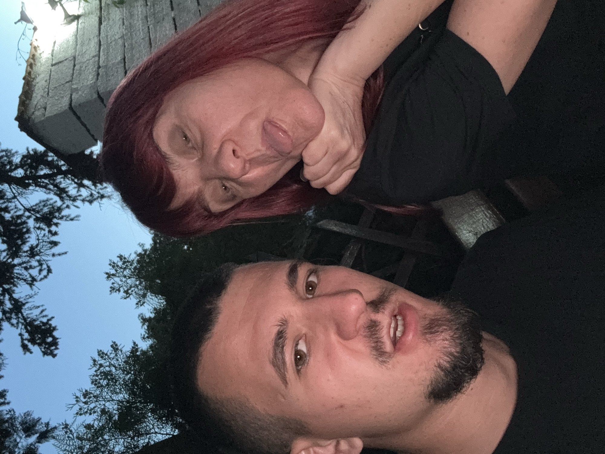 A selfie taken at night features two individuals. The woman, with reddish-purple hair, is positioned above and to the left of the man. She is making a face with her tongue sticking out and her hand clasped around her neck. The man below her has dark hair and a beard, and he is looking upwards at the camera. Both are wearing dark clothing. Behind the man, trees and the evening sky are visible. Behind the woman, a structure with stacked stone blocks is partially visible. Based on the stone structure, the location appears to be near the Ancient Serdica Archaeological Complex in Sofia, Bulgaria.