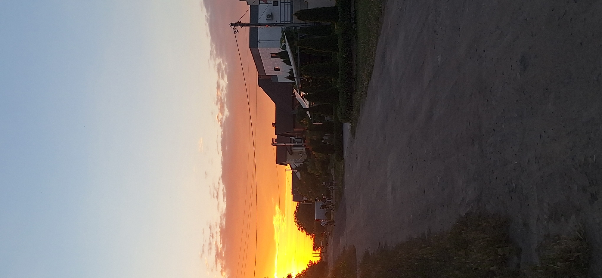 The image depicts a dirt road in a residential area during sunset. The sky is a gradient of orange and blue, with scattered clouds. On the right side of the road are several houses and manicured evergreen trees. On the left side of the road, there is a grassy verge and more trees. In the distance, a white van and several people on bicycles are visible on the road. The overall scene suggests a tranquil evening in a suburban or village setting.