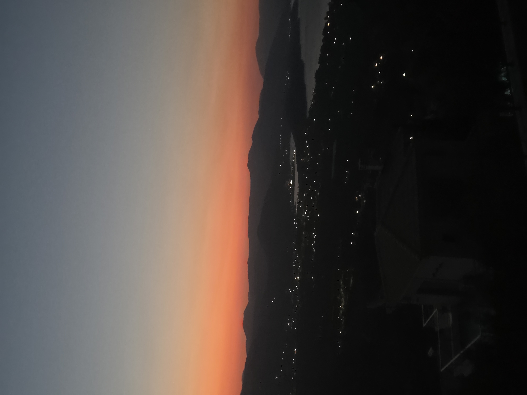 The image depicts a panoramic view of a coastal town at dusk, likely in the Municipal Unit of Asini, Greece, given the presence of rolling hills, a body of water, and scattered lights of the settlement. The sky exhibits a gradient of colors, transitioning from a deep blue at the top to a vibrant orange and pink hue near the horizon, indicating the sunset. Silhouetted mountains form the backdrop, with their contours softened by the twilight. Below, the town lights twinkle like scattered diamonds, suggesting a populated area nestled amongst the hills and along the coastline. A body of water, possibly a bay or lake, is visible to the right, reflecting the dimming light. In the foreground, the dark silhouette of a building with a tiled roof and a terrace is prominent, partially obscuring the view of the town. The overall scene conveys a sense of tranquility and the beauty of a Mediterranean landscape at the end of the day.