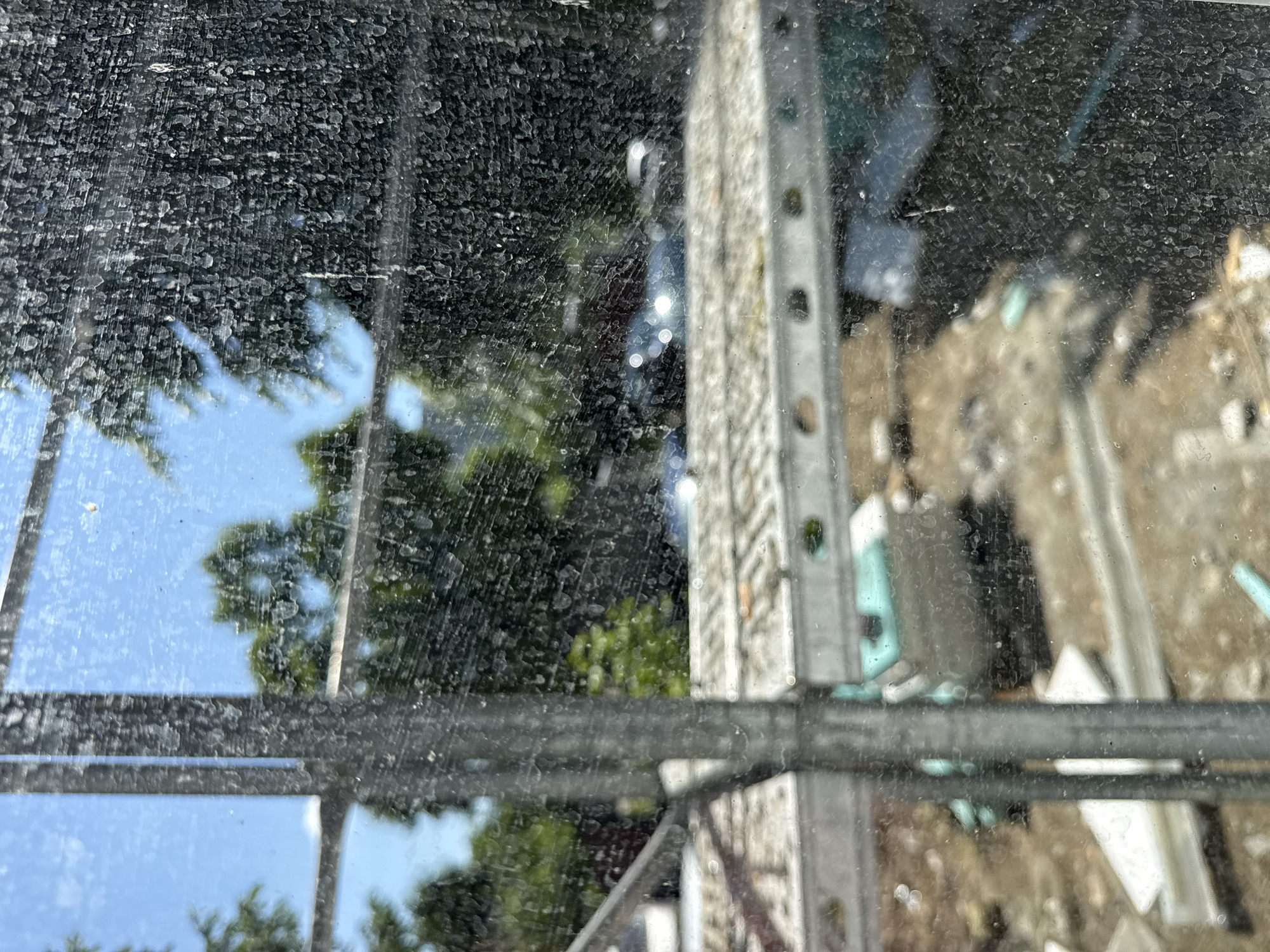 The image appears to be taken through a dirty or dusty window. The window has a metal frame and grid pattern. Through the window, there's a view of a construction site or demolition area. Scaffolding is visible, along with debris and building materials scattered on the ground. Vegetation, likely trees, and a glimpse of blue sky are visible in the background. The location is consistent with an outdoor scene, potentially in the city of Lovech, Bulgaria, where construction or demolition work is taking place.