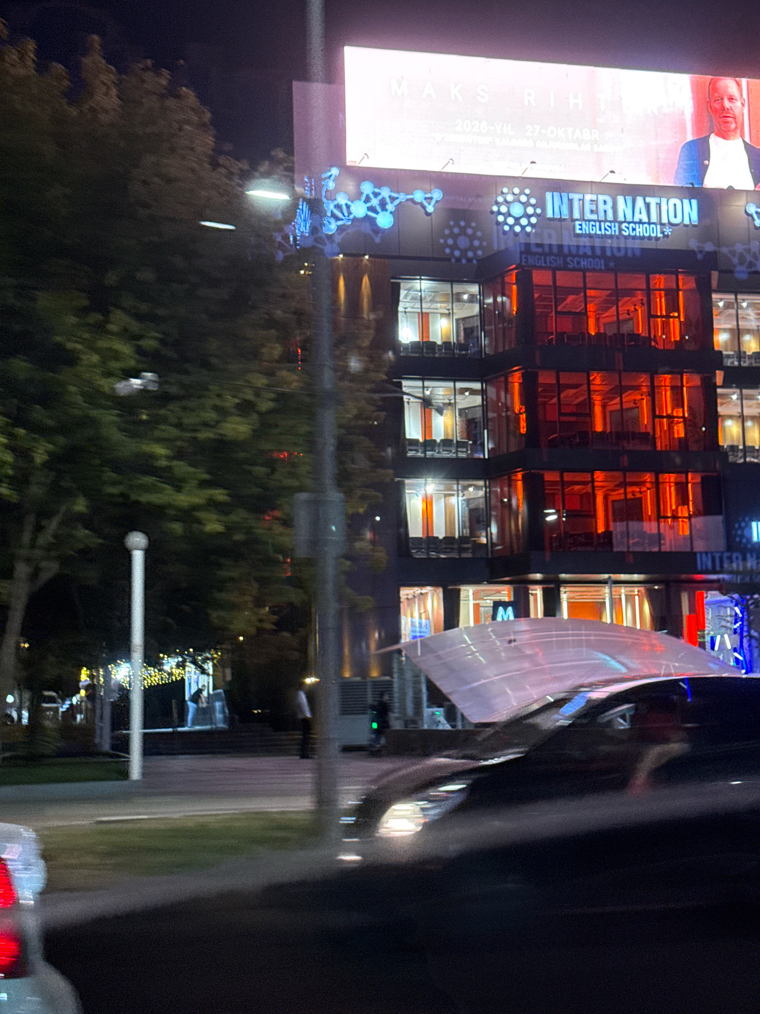 The image shows a night view of the "Inter Nation English School" in Tashkent, Uzbekistan. A large billboard with the text "MAKS RIHT" and a man's face is visible at the top of the building. The building itself has many windows illuminated with red and orange light. In the foreground, a car with its hood open is parked in front of the school. The street is lit by streetlights and decorative lights. Trees are visible on the left side of the image.