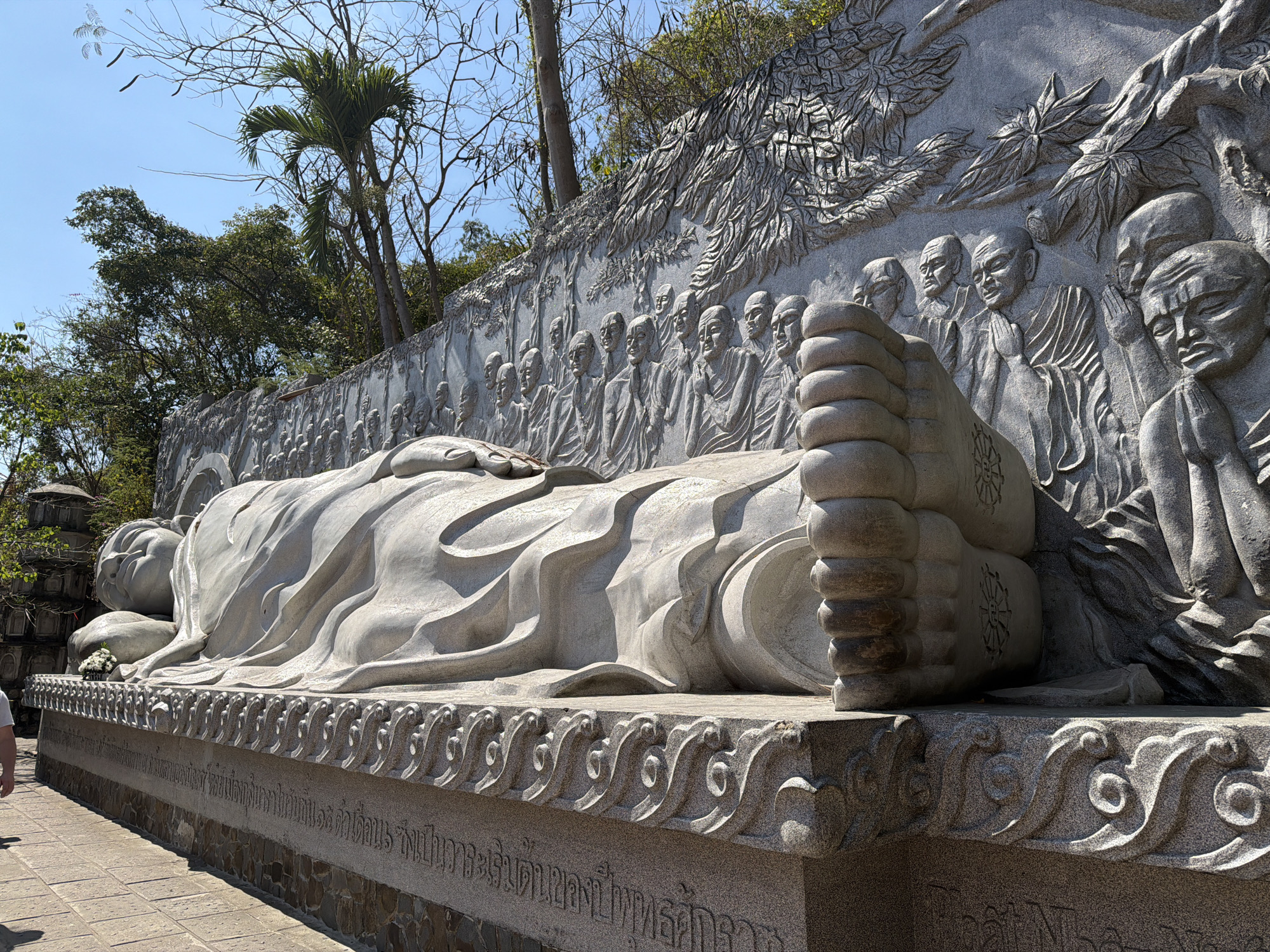 The image shows a large, intricately carved stone statue of a reclining Buddha at the Long Son Pagoda in Nha Trang Ward, Vietnam. The Buddha is depicted lying on his side, draped in flowing robes. Behind the statue is a relief carving depicting a procession of monks and foliage. The base of the statue is decorated with a carved wave pattern and inscriptions in Vietnamese script. Above the monument are trees, including palm trees, set against a clear blue sky. The scene appears to be outdoors, with sunlight illuminating the sculpture. Part of a person is seen in the lower left corner of the picture.