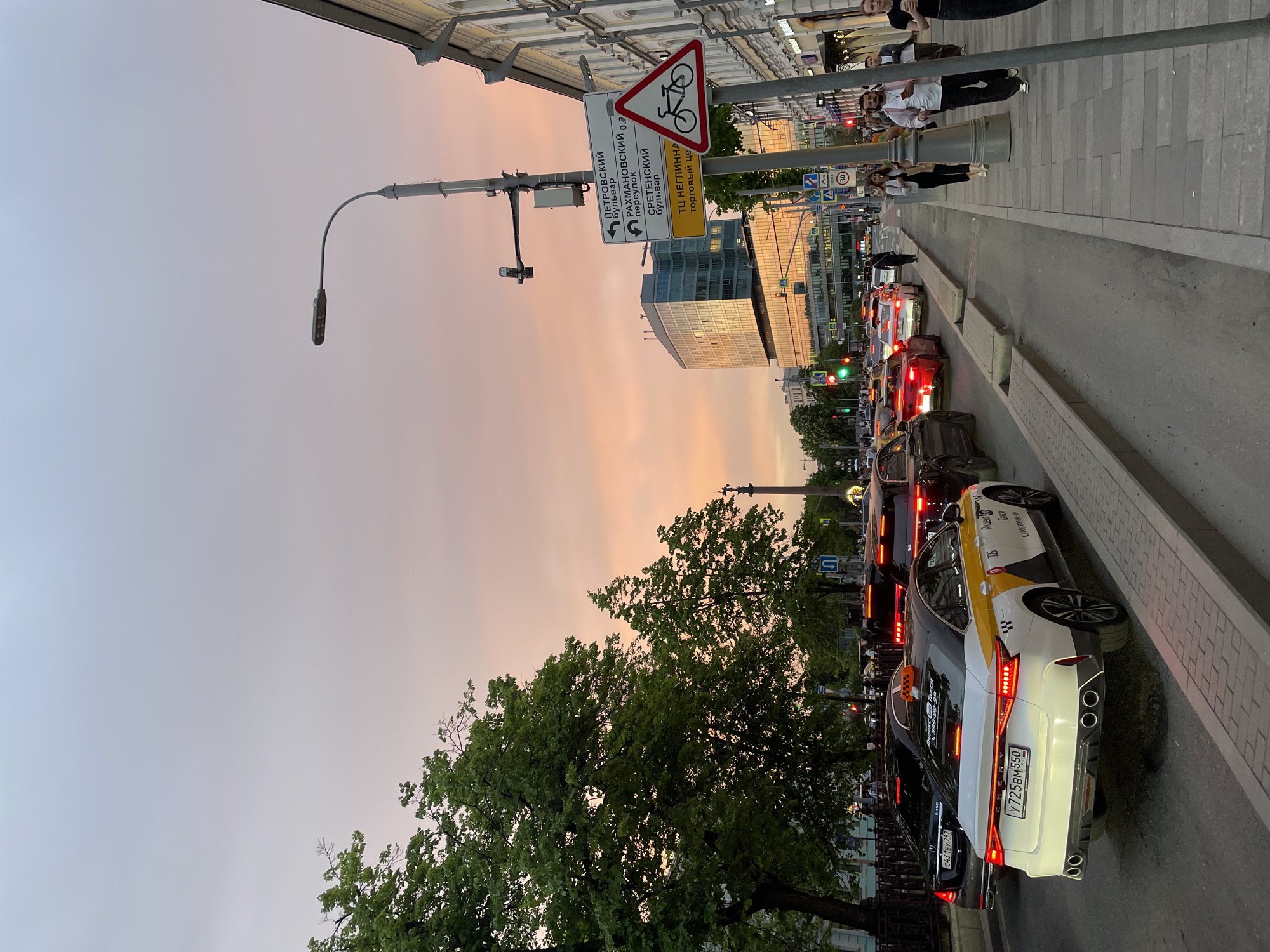 The image captures a street scene in Moscow, Russia, at dusk or twilight. The sky exhibits a gradient of colors, from light gray to pink, suggesting either sunrise or sunset. A row of stationary cars, possibly in a traffic jam, lines the right side of the street. At least two of the cars are identifiable as taxis, indicated by the checkerboard patterns and roof signs. License plates are visible on the cars, and the characters appear Cyrillic. A green light is visible in the distance. On the left side of the image is a large tree with green leaves. A street lamp and various road signs are present, including one depicting a bicycle lane and street name signs in Cyrillic. The buildings in the background reflect the colors of the sky. People are walking on the sidewalk to the right. The location is identifiable as Moscow by the license plates and Cyrillic text.