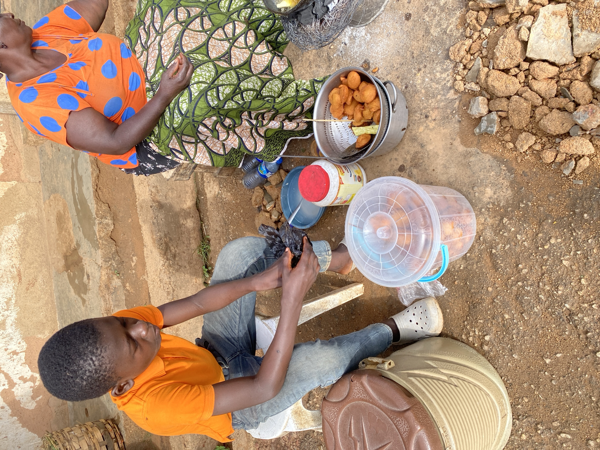 A young Black boy wearing an orange polo shirt and blue jeans sits on a white stool, holding a black plastic bag filled with food items. He is looking down at the bag. Beside him is a large, light brown plastic container with a textured lid. To his right, a woman wearing an orange top with blue polka dots and a green and white patterned skirt sits, her hands engaged with food preparation. In front of the woman, a metal colander filled with fried dough balls rests on top of a silver pot. A clear plastic bucket containing more fried dough is positioned to the left of the colander. The scene appears to be outdoors on a dirt ground with scattered rocks and dried leaves. Behind the individuals, a rough, textured wall and steps are visible. The environment suggests a street-side food vending setup. The presence of both individuals, the food preparation items, and the outdoor setting strongly indicate a commercial activity, likely a food stall. The overall atmosphere is that of a busy, working environment within a Nigerian city.
