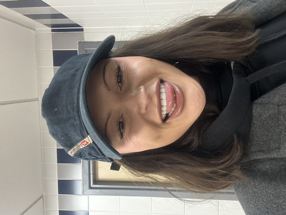 A woman smiles, showcasing her teeth. She wears a dark-colored cap with a visible brand label. She is dressed in a black hoodie and a gray coat. Her hair is shoulder-length and brown. The background features a tiled wall, with alternating rows of white and blue-and-white tiles. A wooden-framed mirror or picture is also visible in the background.