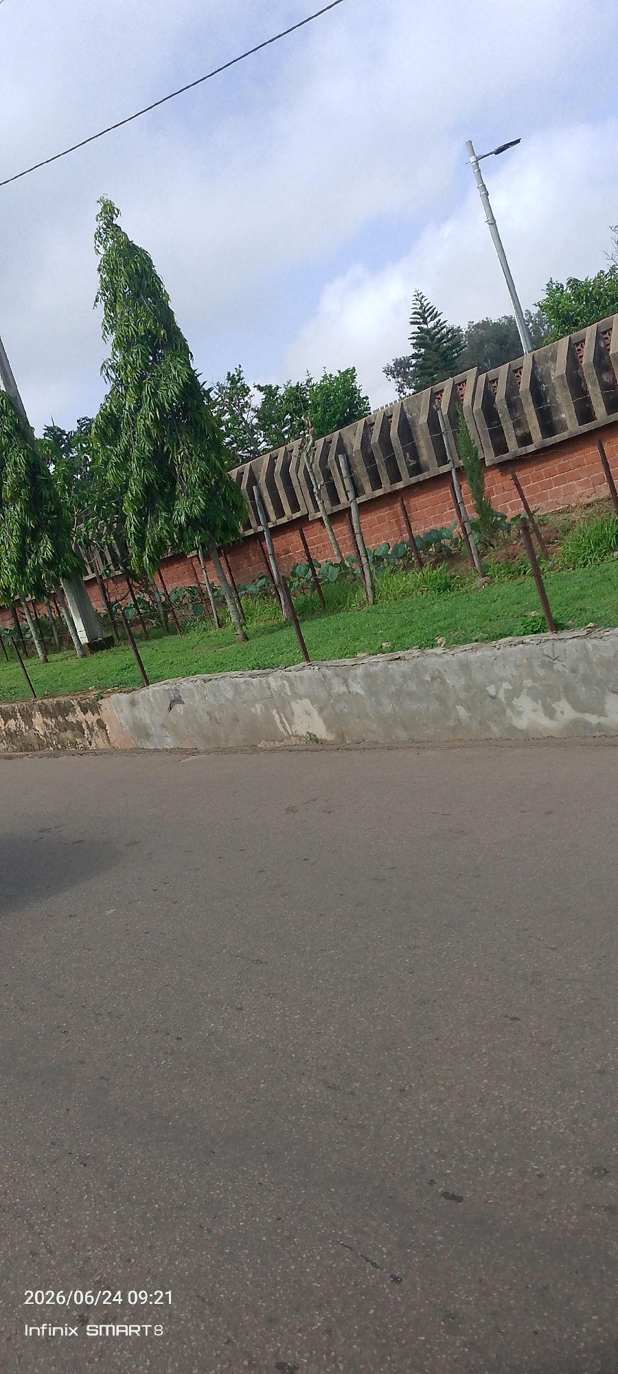 The image depicts a paved road in Jos, Nigeria, on a cloudy day. The road occupies the lower half of the frame, with a textured grey surface. A concrete barrier, showing signs of age and some wear, runs along the edge of the road. Behind the barrier is a strip of green grass, and beyond that, a garden area with some low-lying plants. A brick wall with a decorative concrete coping runs along the back of the garden. Several tall, slender trees with dense green foliage line the area in front of the wall, casting thin shadows on the grass. In the background, the sky is overcast with large, diffused clouds. A lamppost is visible on the right side, above the wall. The overall scene suggests an outdoor setting, possibly a roadside or an area adjacent to a property. No people are visible.
The bottom left corner of the image contains text indicating the date and time: "2026/06/24 09:21" and "Infinix SMART 8", likely a timestamp and the device used to capture the photo. The time of day is morning, as indicated by the text, and the weather appears to be cloudy. The environment suggests a typical urban or suburban landscape in Nigeria.