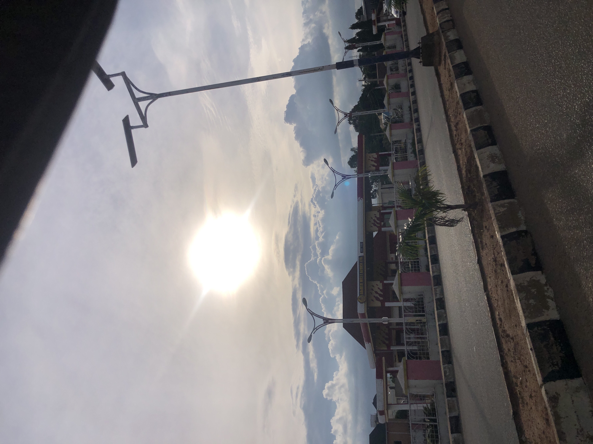 The image depicts a street scene in Jos, Nigeria, under a bright, partly cloudy sky. The sun is prominently visible, casting a strong glare. A gas station, identifiable by its red and white architecture and visible signage, is situated in the background. Several streetlights are present, with one particularly large, modern-looking streetlight with two light fixtures dominating the right side of the frame. A smaller, ornamental streetlight is visible in front of the gas station. The road in the foreground is paved, with a curb and a dirt median. A small palm tree grows in the median. The overall impression is of a typical urban street on a sunny day.