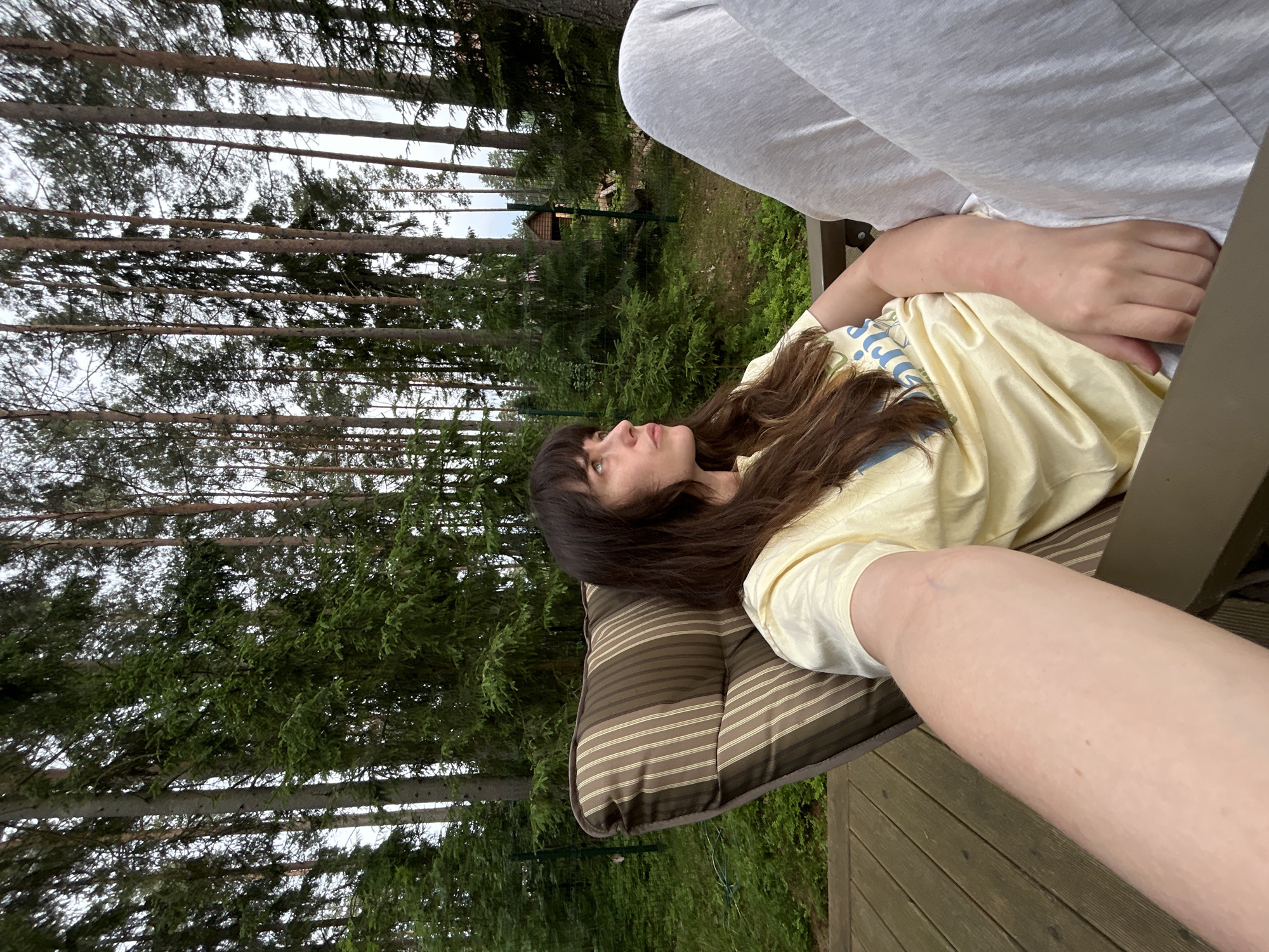 In Shepelyovo, Russia, a woman with long, dark hair sits in a striped patio chair on a wooden deck. She is wearing a light yellow t-shirt and light gray sweatpants. The woman is looking off to her right, her head tilted upwards. The background features a dense forest with tall evergreen trees. A fence runs along the edge of the property, with a small wooden structure visible in the distance. The overall scene suggests a relaxed outdoor setting.