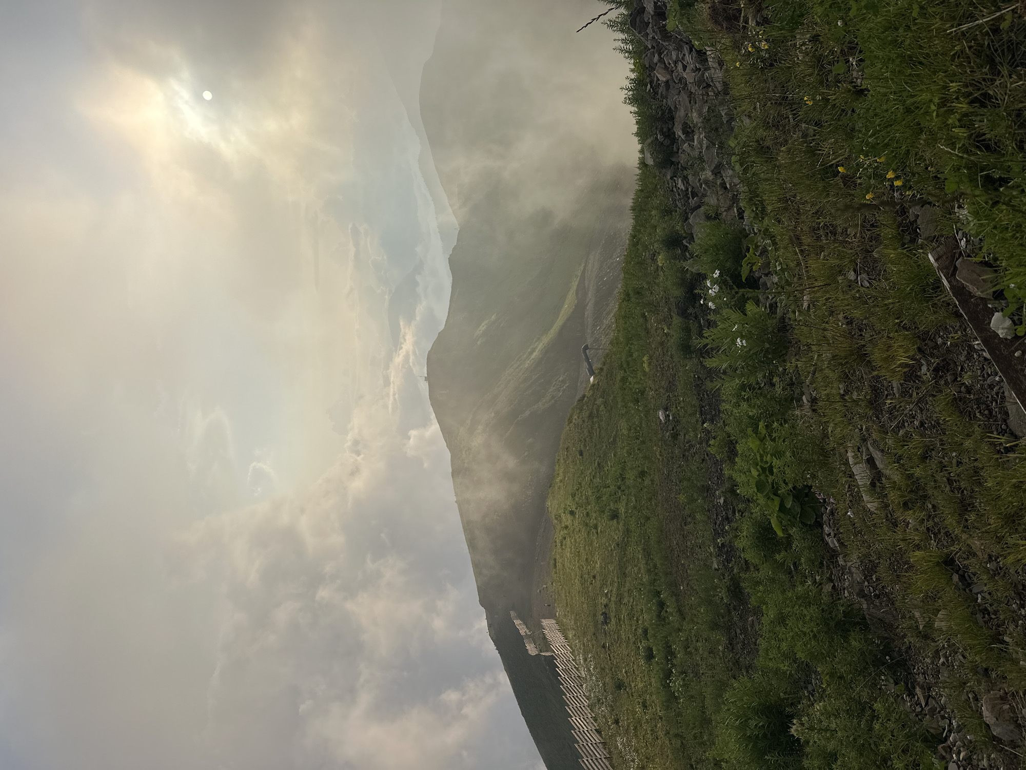 The image depicts a mountainous landscape with a winding dirt path and a wooden fence along its edge, likely a hiking trail or ski slope in Estosadok, Russia. The sun is partially obscured by clouds, casting a diffused light over the scene. Mist or fog partially envelops the mountain peaks in the background, adding a sense of depth and mystery. The foreground is covered with green grass and scattered rocks, with a few wildflowers adding pops of color. The overall atmosphere is serene and natural, highlighting the beauty of the mountain environment. There are no visible people or specific objects that indicate a particular event or activity.