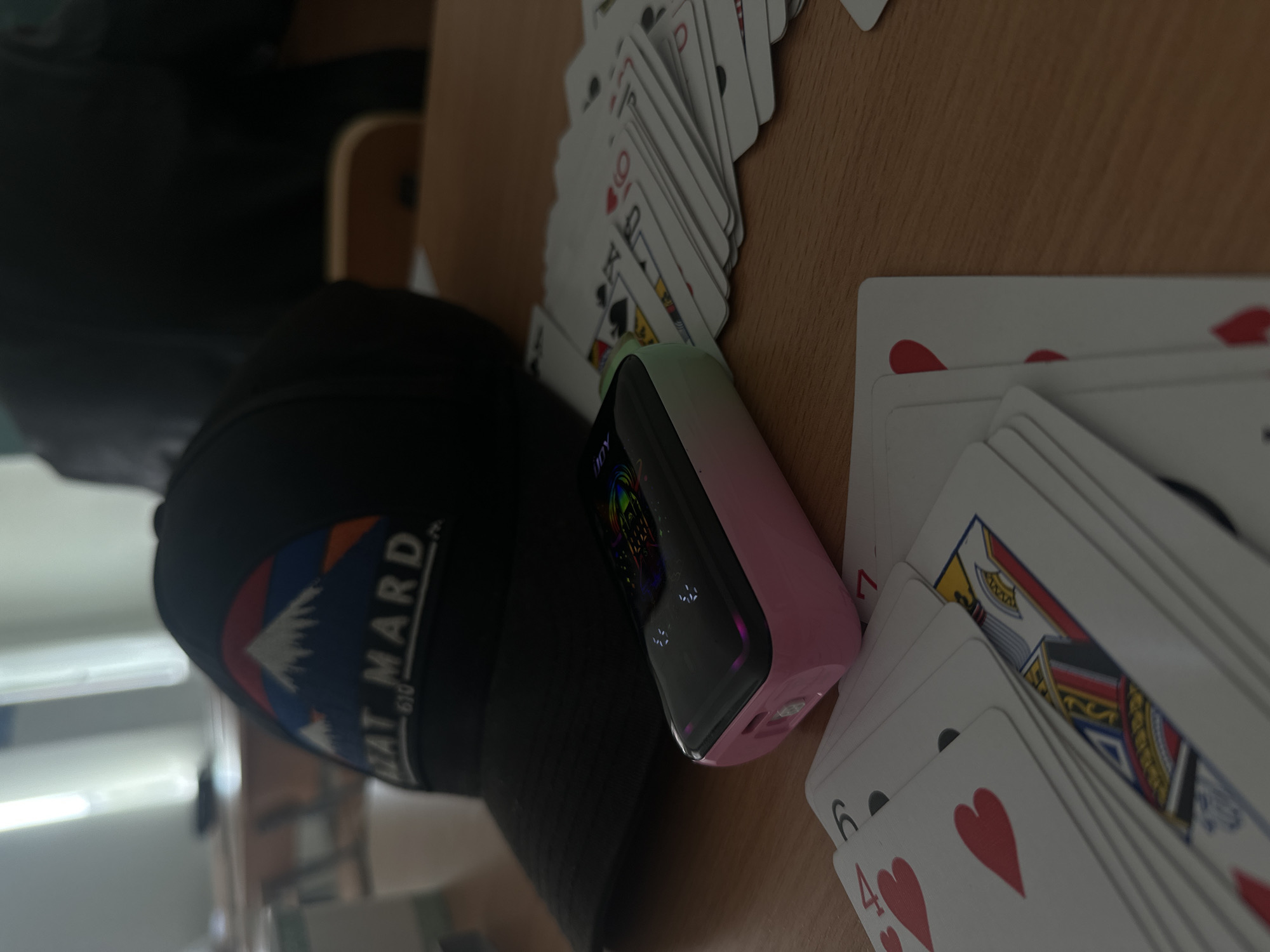 The image, captured from a low angle and rotated to a portrait orientation, displays a collection of items on a light brown wooden surface, likely a table or desk, indoors.
The primary subjects include several spreads of playing cards, a black baseball cap, and a distinct electronic device. Two main groups of playing cards are visible. To the upper left, a fanned arrangement of cards shows a mix of suits and ranks, including spades and clubs, with visible Kings (K), Queens (Q), and tens (10). To the lower right, another stack of cards is fanned out, primarily showing hearts, with visible numbers such as 4, 6, and 7.
Nestled between the cards and the cap is a sleek, rectangular electronic device. It features a gradient color scheme, transitioning from a light pastel green at one end to a soft pink at the other. The device has a dark, reflective screen that displays colorful digital graphics and some text, including the word "JOY" and small white indicators, possibly battery levels or settings.
To the left of the electronic device is a black baseball cap. It features a prominent patch with a mountain range motif in shades of white, blue, orange, and green. Below the mountains, white text reads "VAT MARD" and "610," with some faint additional text below that.
The background is softly blurred, showing hints of indoor furniture and walls, suggesting a casual setting. The overall impression is one of leisure, perhaps a break from an activity like playing cards. No specific time of day or emotions are discernible.