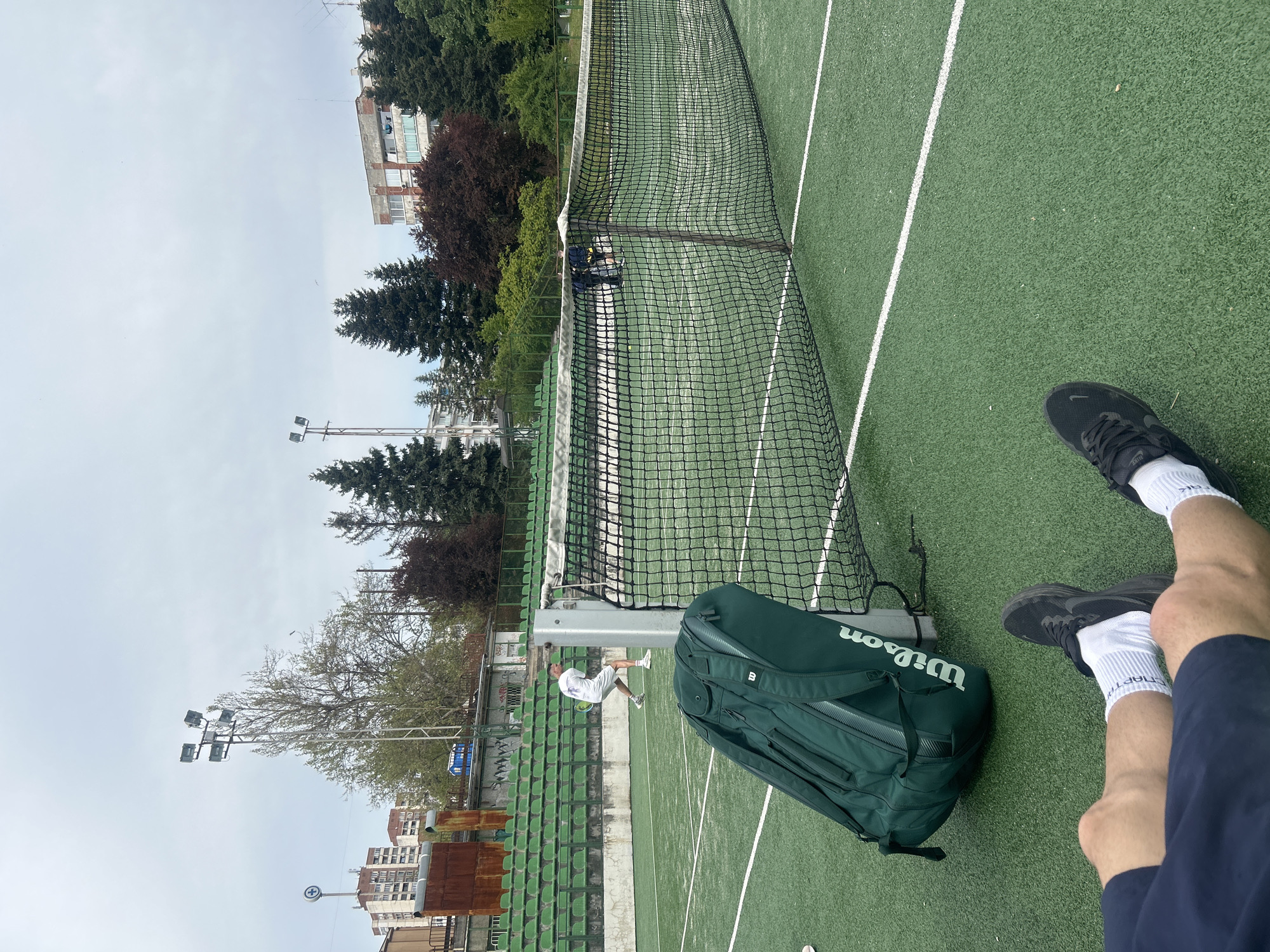 The image shows a person sitting on a green tennis court, with their legs and feet visible. They are wearing black sneakers and white socks, and navy blue shorts. Next to them is a green Wilson tennis bag. In the background, there is a tennis net, a tennis player, and green bleacher-style seating. The setting appears to be an outdoor tennis court in Veliko Tarnovo, Bulgaria, as visible in the background are buildings and lighting fixtures that are similar to those of the city.