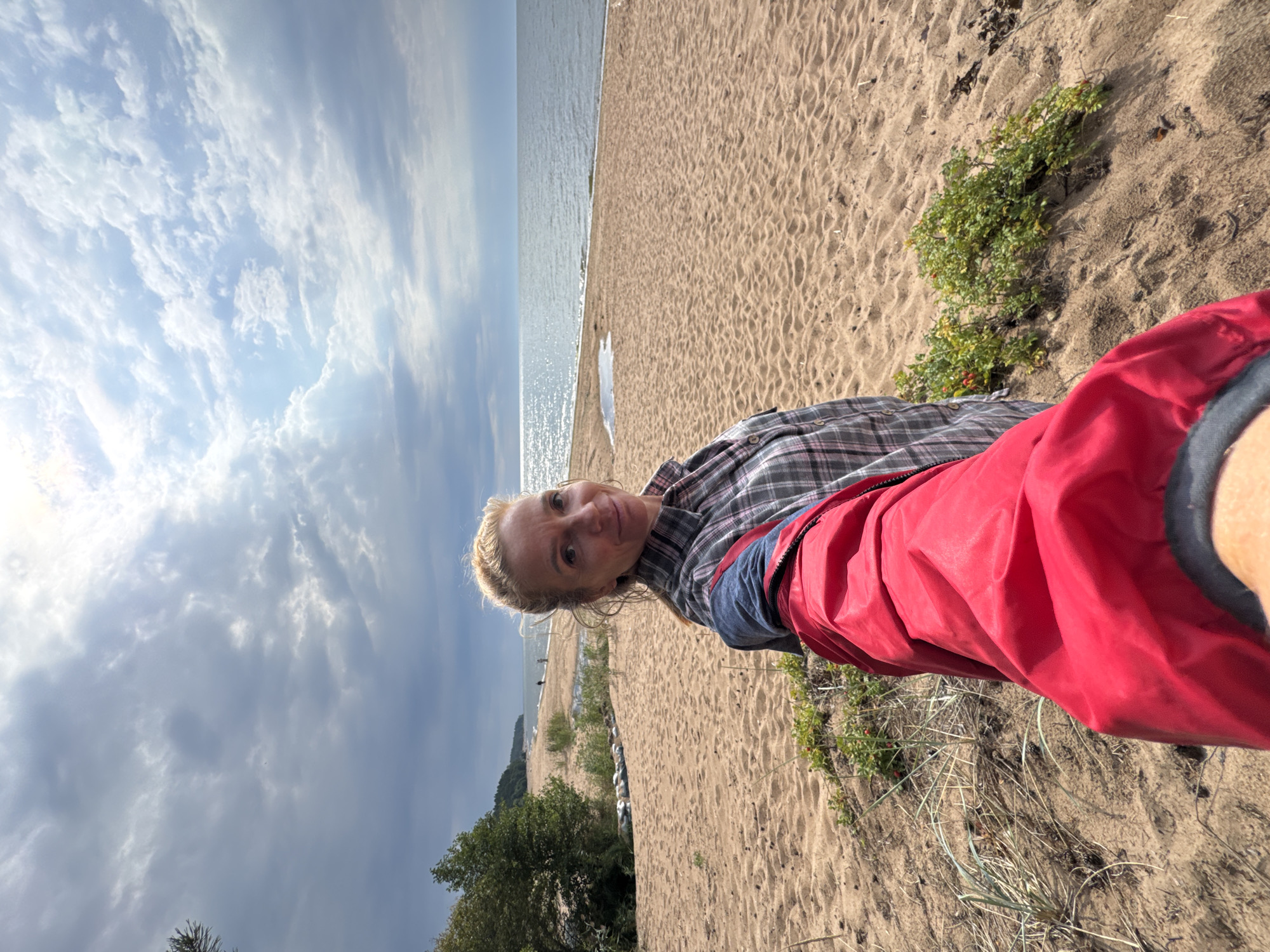 A woman with blonde hair pulled back is taking a selfie on a sandy beach. She is wearing a plaid shirt and a red jacket. The beach is lined with vegetation and trees, and the sea stretches out to the horizon under a partly cloudy sky. The location is identified as Zelenaya Roshcha, Russia.