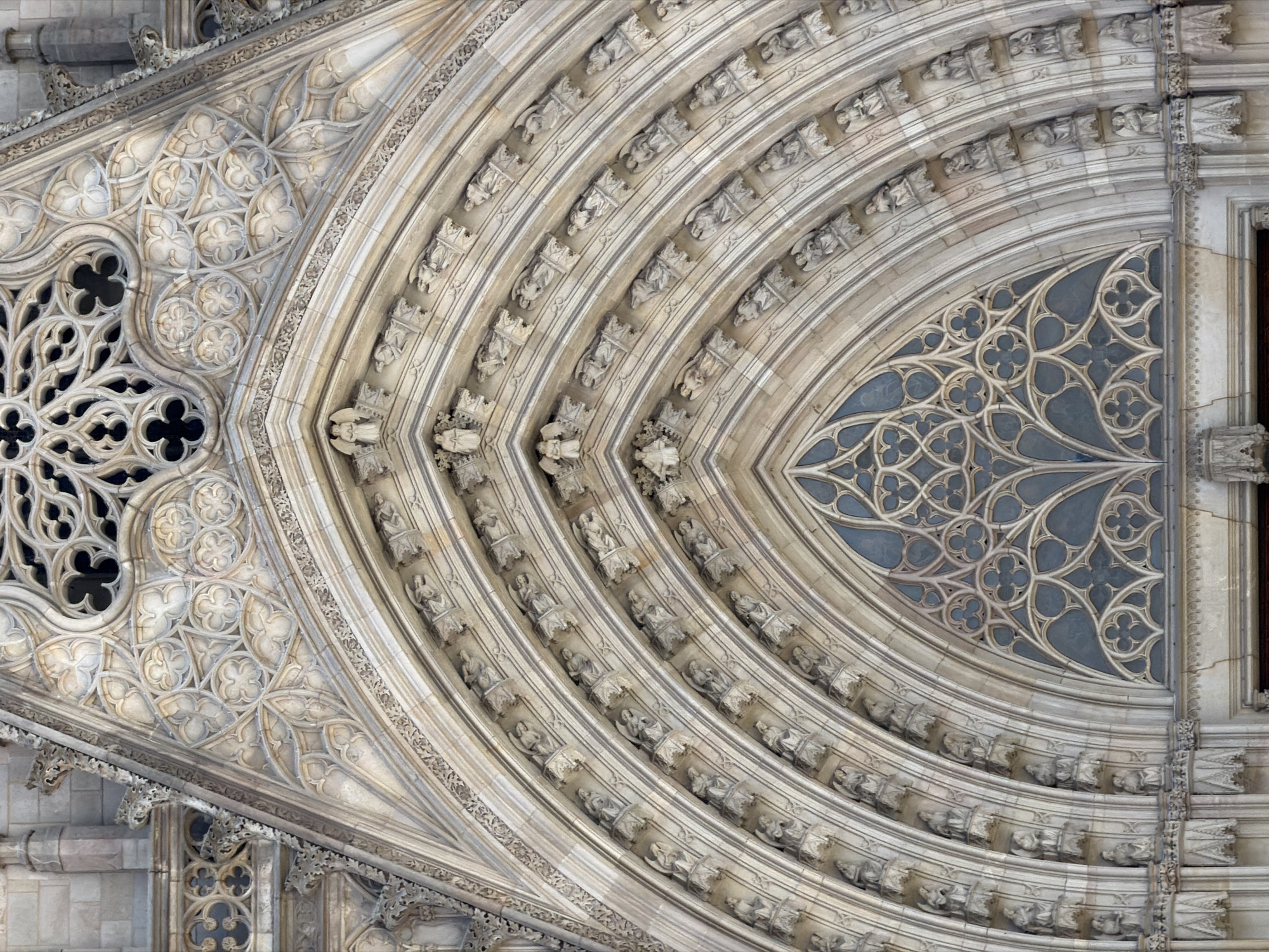 This image is a close-up, vertical shot of the exterior facade of a large Gothic cathedral, likely the Barcelona Cathedral in Barcelona, Spain, given the architectural style and prominent rose window. The ornate stonework features multiple layers of pointed arches, characteristic of Gothic architecture. Each arch is adorned with repeating decorative elements, including sculpted figures, possibly saints or angels, positioned within small niches.
The central focus of the image is a large rose window, a circular window divided into intricate tracery patterns, with a dark, stained-glass interior. Above and around the rose window, the stonework is heavily carved with floral motifs and geometric patterns, forming a complex decorative scheme. The material appears to be weathered stone, exhibiting varying shades of beige and gray, suggesting age and exposure to the elements. The lighting indicates daylight, casting subtle shadows that highlight the three-dimensional nature of the carvings. No people are visible in the frame. The perspective is from below, looking upwards towards the architectural details.