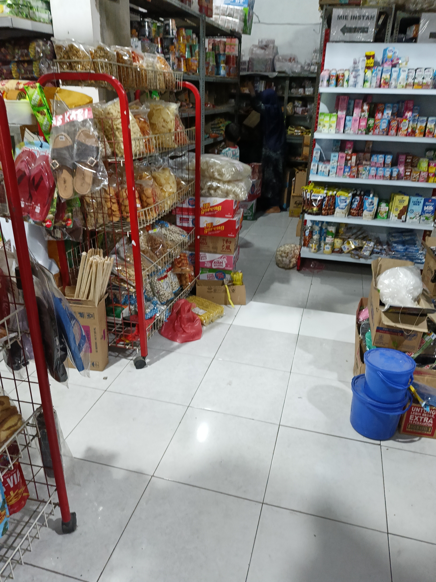 The image depicts the interior of a small convenience store, likely in Sukorini, Indonesia. The store is packed with various goods, predominantly food items and household products. In the foreground, a red metal rack displays packages of snacks, sandals, and what appear to be breadsticks. To the right, shelves are laden with bottled drinks, likely milk and juice, and boxed food items. A person in dark clothing, possibly a store employee, is visible in the background interacting with the shelves. The floor is tiled with a light-colored pattern. Several blue buckets are stacked near the right side of the frame, in front of a shelf containing boxed goods. The overall impression is one of a modest, well-stocked retail space.