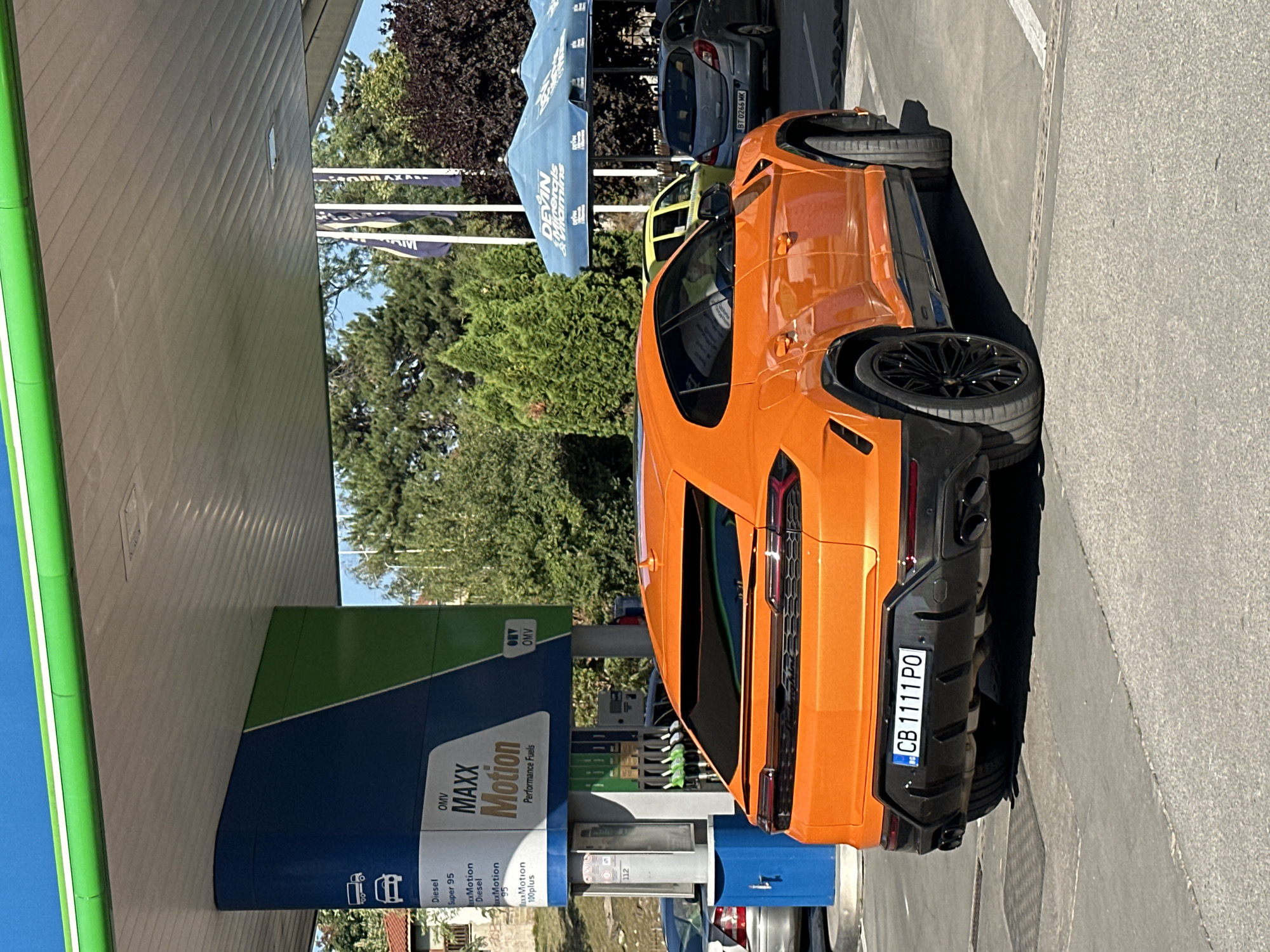 This is a vertically oriented, slightly blurry photograph taken outdoors during the daytime, likely on a sunny day given the strong shadows. The main subject is a bright orange sports car, possibly a Lamborghini Urus, parked at a gas station. The gas station has green and blue branding with the text "OMV MAXX Motion Performance Fuels" visible. There are several gas pumps with green nozzles. In the background, trees and a blue canopy are visible, along with other cars. One car in the background has the license plate "BT 0266 MK". The orange car has a license plate that reads "CB 1111 P0". The scene appears to be a refueling stop.