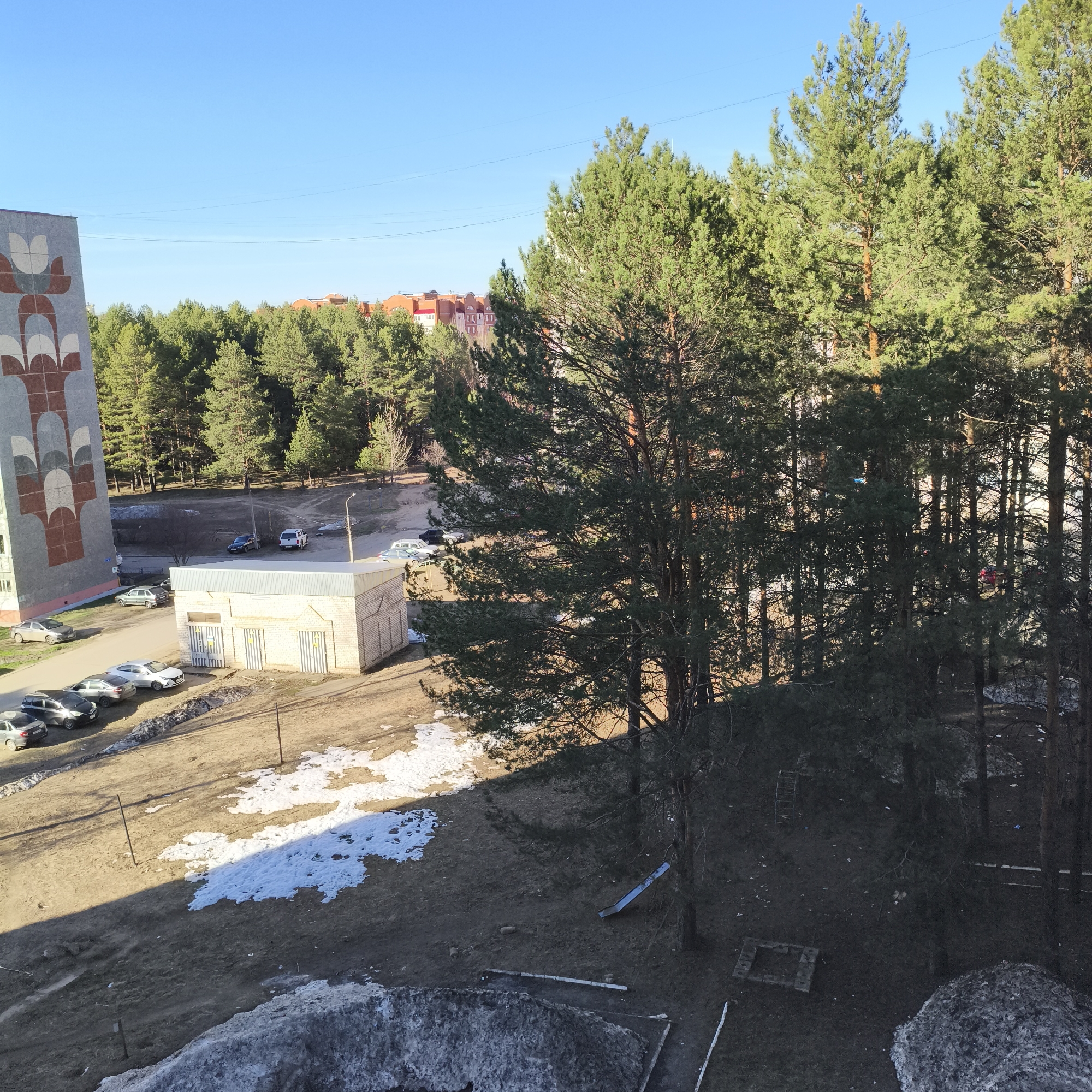 This image depicts an outdoor scene in Chaikovsky, Russia, likely during early spring. The time of day appears to be either morning or late afternoon, indicated by the bright sunlight and prominent shadows. The sky is clear and blue.
On the left, a tall, multi-story residential building is visible, featuring a large, distinctive decorative mural on its side with geometric patterns in shades of grey, brown, and off-white, resembling stylized floral motifs. Below this building, several cars are parked on an unpaved area.
In the middle ground, a single-story brick building, possibly containing garages or utilities, stands with multiple striped metal doors. A street lamp post is situated nearby. The ground in this area consists of bare earth with patches of melting, dirty snow.
The right side and background of the image are densely covered by a forest of tall pine trees, their green needles contrasting with the clear sky. In the far distance, beyond the treeline, residential buildings with reddish-brown roofs are visible. Within the shadowed areas under the trees, a small metal slide and a rectangular concrete or brick outline are discernible on the ground. A large, irregular mound of what appears to be compacted snow or ice occupies the lower right foreground. There are no people visible, and no specific activities or events are taking place. No visible text is present in the image.