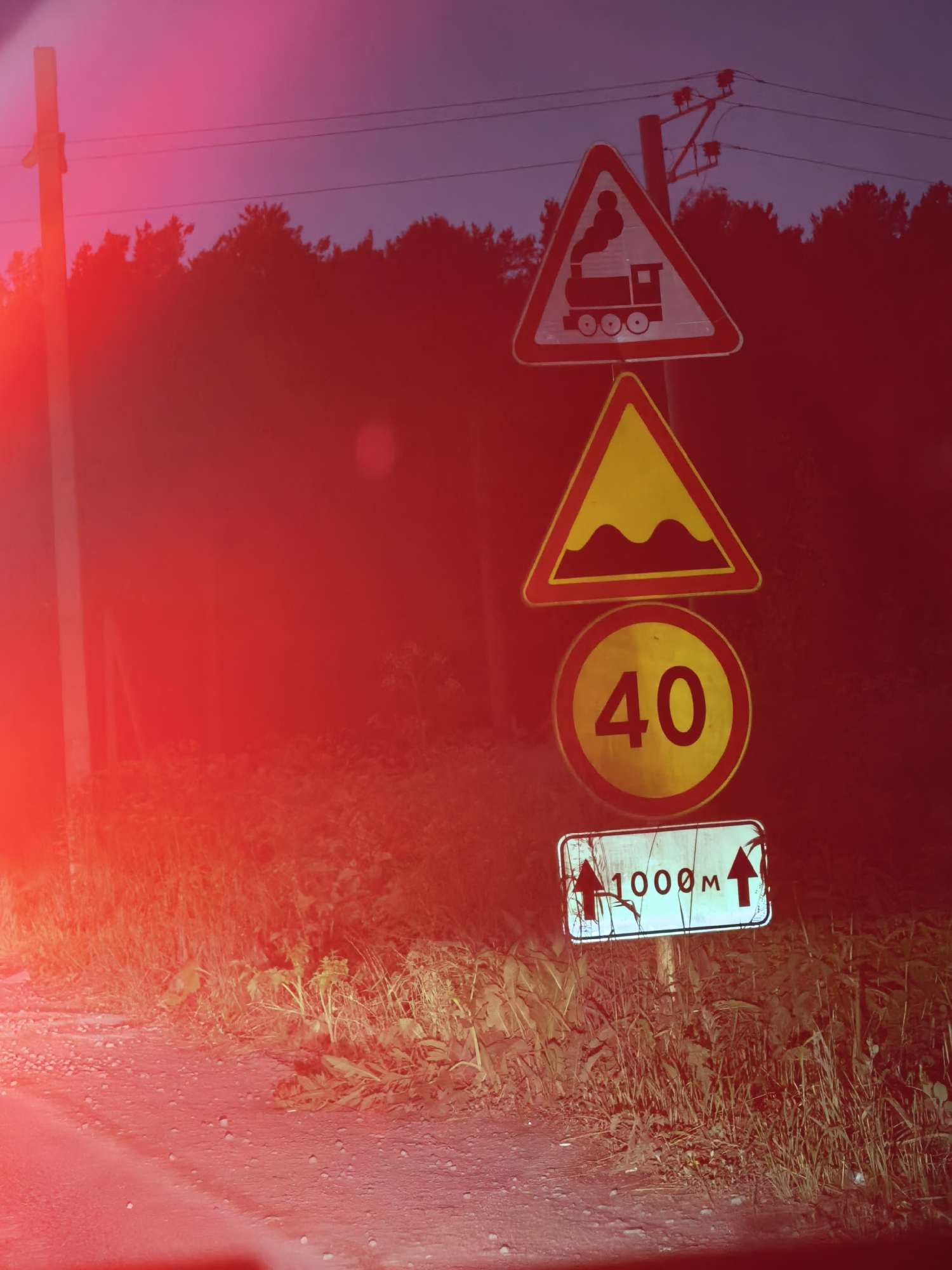 The image shows a series of traffic signs on a pole at dusk or night, with a strong red light illuminating the foreground and part of the scene. The signs are stacked vertically. The top sign is a red triangle with a black silhouette of a steam locomotive, indicating a railroad crossing. Below that is a yellow triangle with a red border, depicting a wavy line that signifies a series of bumps or an uneven road surface. The next sign is a circular speed limit sign with a red border and a yellow interior, displaying the number "40". The bottom sign is a rectangular white sign with a red border, featuring two black upward-pointing arrows flanking the text "1000 m". The background consists of silhouetted trees against a dark sky with visible power lines and utility poles. The ground in the foreground is a gravelly road surface with some vegetation along the side. The intense red light appears to be from a vehicle's taillight or emergency light, casting a strong glare and obscuring some details of the environment.