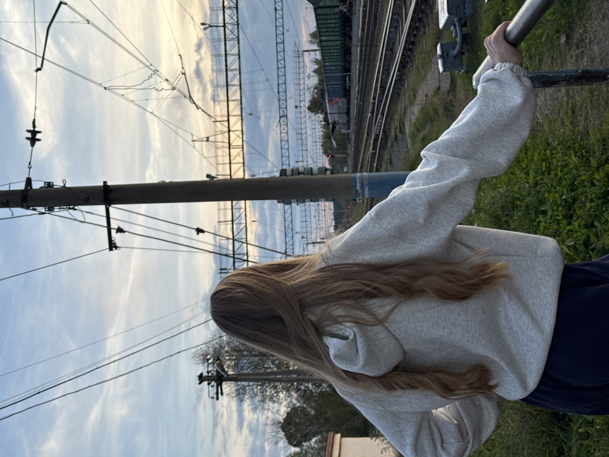 A young woman with long, blonde hair is standing with her back to the camera, looking out at a train station. She is wearing a light grey hooded sweatshirt and dark blue pants. Her arms are resting on a metal railing in the foreground. In the background, train tracks stretch into the distance, with several train cars stopped on one of them. Overhead, a complex network of electrical wires and poles crisscrosses the sky, which is a pale blue with scattered clouds and the warm glow of a setting sun. The area around the tracks is covered in green grass and some trees can be seen in the distance. The overall scene suggests an outdoor, rural or semi-rural location, consistent with the outskirts of a settlement like Komarihinskiy, Russia, likely during the late afternoon or early evening.