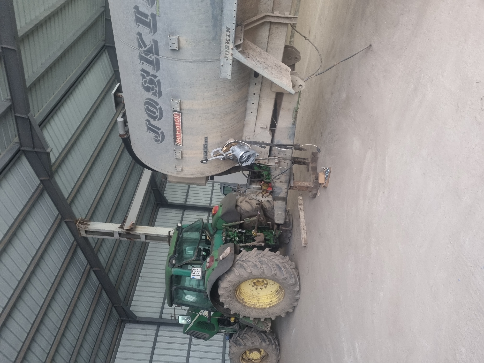 The image shows a green John Deere tractor coupled to a silver Joskin agricultural tanker inside a building with a metal frame and corrugated metal siding. The tractor has the license plate "1.XEX 1741," suggesting it's registered in Bulgaria. The scene is likely in a rural or agricultural setting. The location could be Ovcharovo, Bulgaria, as requested, but there are no specific location markers within the image to confirm.