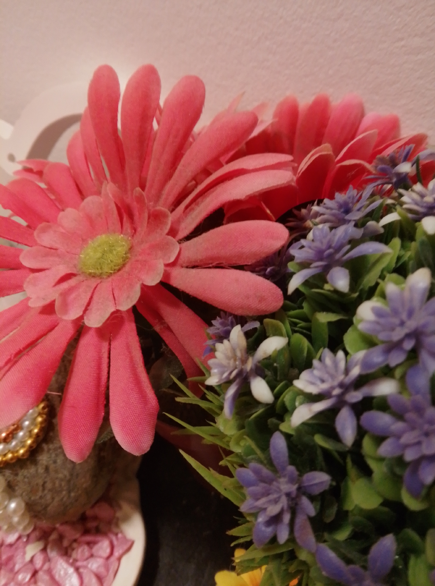 This is a close-up, eye-level, indoor photograph of artificial flowers. In the foreground, a large, bright pink daisy-like flower dominates the left side of the frame. Its petals are broad and textured, with a fuzzy yellow-green center. To the right of the pink flower, a cluster of smaller, delicate purple flowers with green foliage is visible. Some of these purple flowers appear to be in various stages of bloom, with some buds and fully opened blossoms. The background is softly blurred, showing a pale pink textured wall or surface. There is also a hint of a white object, possibly a decorative element, in the upper left corner. The lighting is soft and diffused, creating a gentle ambiance. The image is focused on the textures and colors of the artificial floral arrangement, highlighting the details of the petals and leaves.