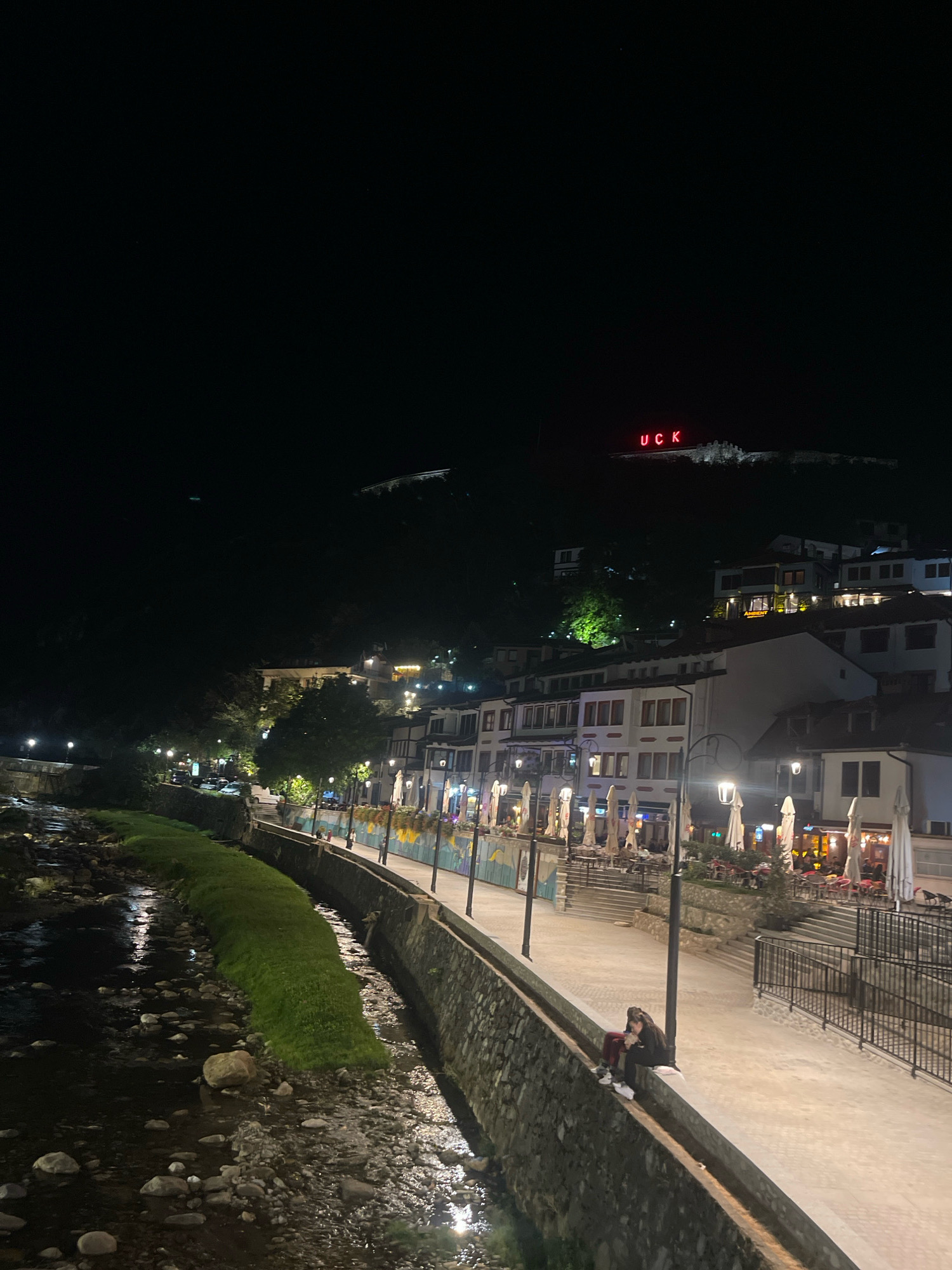The image shows a nighttime view of the Lumbardhi River in Prizren, Kosovo. Buildings with illuminated windows line the river on the right side, and outdoor seating areas with umbrellas are visible. A stone wall and a sidewalk run alongside the river. Two individuals are seated on the stone wall overlooking the river. In the background, high on a hill, the red neon sign "UCK" is illuminated. The riverbed is visible with rocks and water. Streetlights illuminate the walkway.