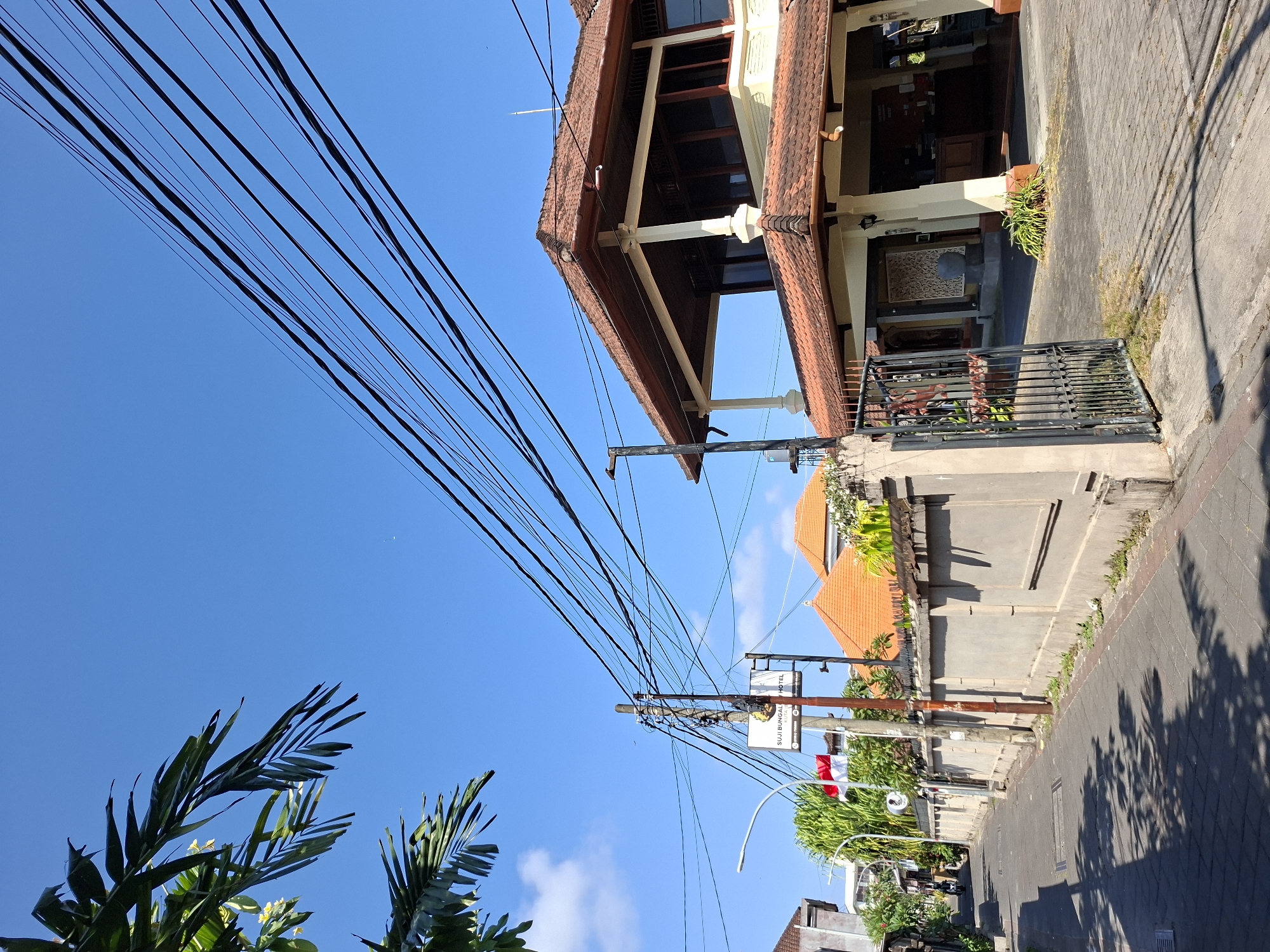 The image depicts a street scene in Kuta, Indonesia, under a clear blue sky. Power lines dominate the upper portion of the frame, strung across the sky. In the foreground, a paved street with cobblestone patterns leads into the distance, flanked by a concrete wall on the right and buildings on the left. Shadows of palm fronds are cast on the street and wall, indicating sunlight.
A building with a traditional Indonesian tiled roof, featuring a large covered porch supported by pillars, is visible on the right. An open gate made of wrought iron provides access to this building's entrance. A sign displaying "SUJI BUNGALOW HOTEL" is mounted on a pole near the street. A red and white Indonesian flag flies on a flagpole behind the sign.
In the distance, a person on a motorcycle is seen riding down the street. Trees and foliage are interspersed throughout the scene, adding to the tropical atmosphere of Kuta. The overall impression is a quiet, sunlit day in a residential or commercial area of the city.
