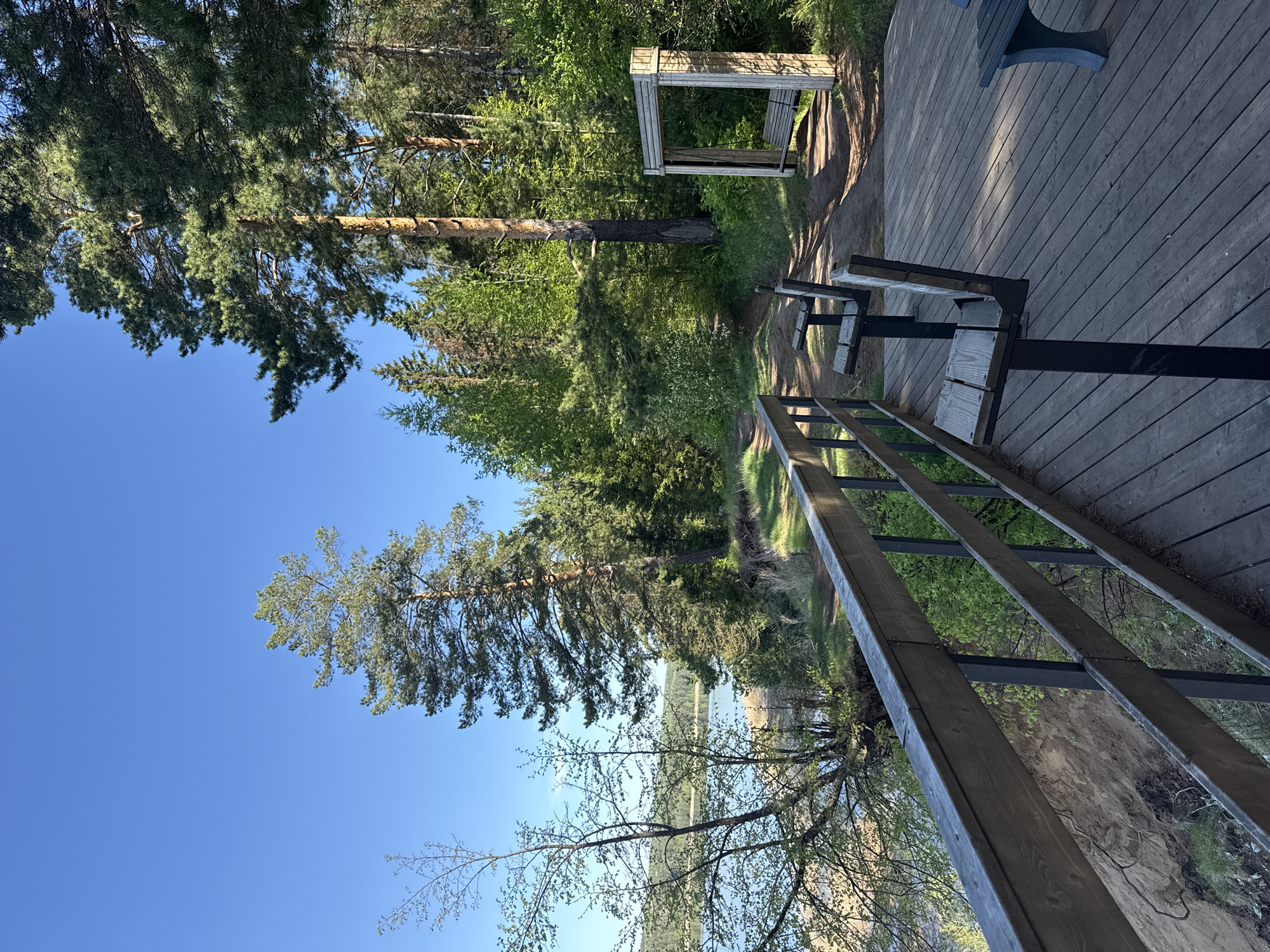 The image shows a view from a wooden deck, possibly near a body of water, with trees and greenery visible. A set of benches is attached to the deck, overlooking the landscape. There is a structure resembling a frame or shelter visible in the distance. The sky is clear and blue. Although the city and country are known, the image doesn't provide any indication of its location being in Votkinsk, Russia.