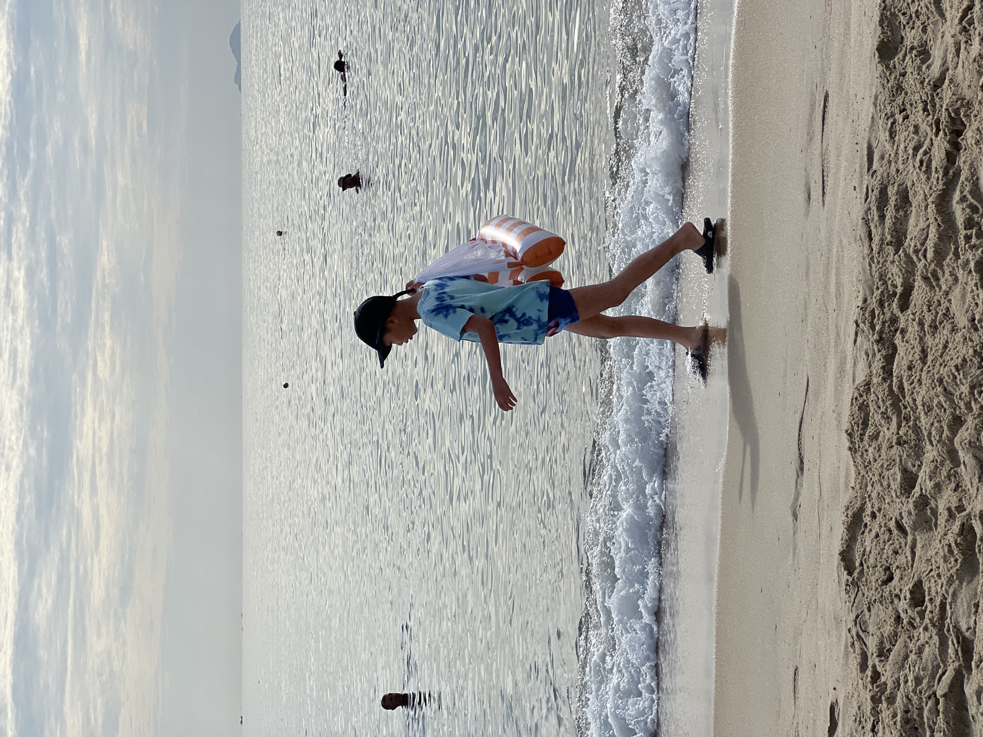 A child wearing a black cap and a blue tie-dye t-shirt walks along the shoreline of a beach in Nha Trang, Vietnam. The child carries a mesh bag containing an orange and white inflatable object. Gentle waves lap the sandy shore. In the background, several people are swimming or wading in the calm sea, with a hazy horizon and a distant island visible. The sky is overcast with scattered clouds.