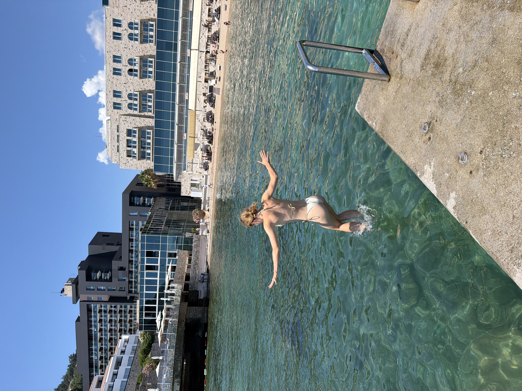In Budva, Montenegro, on a bright, sunny day, a young woman in a white bikini is captured mid-jump into the crystal-clear, turquoise water. She is positioned in the foreground, facing away from the camera with her arms outstretched, her legs bent, and her feet splashing into the sea. The water reveals a rocky seabed, indicating shallow depths. To her right, a concrete pier extends into the water, equipped with a metal ladder for access. In the background, a modern beach resort complex lines the shore, featuring multiple hotel buildings with contemporary architecture and numerous beach umbrellas providing shade for sunbathers on the pebble beach. The sky is a vivid blue, with a few scattered white clouds.