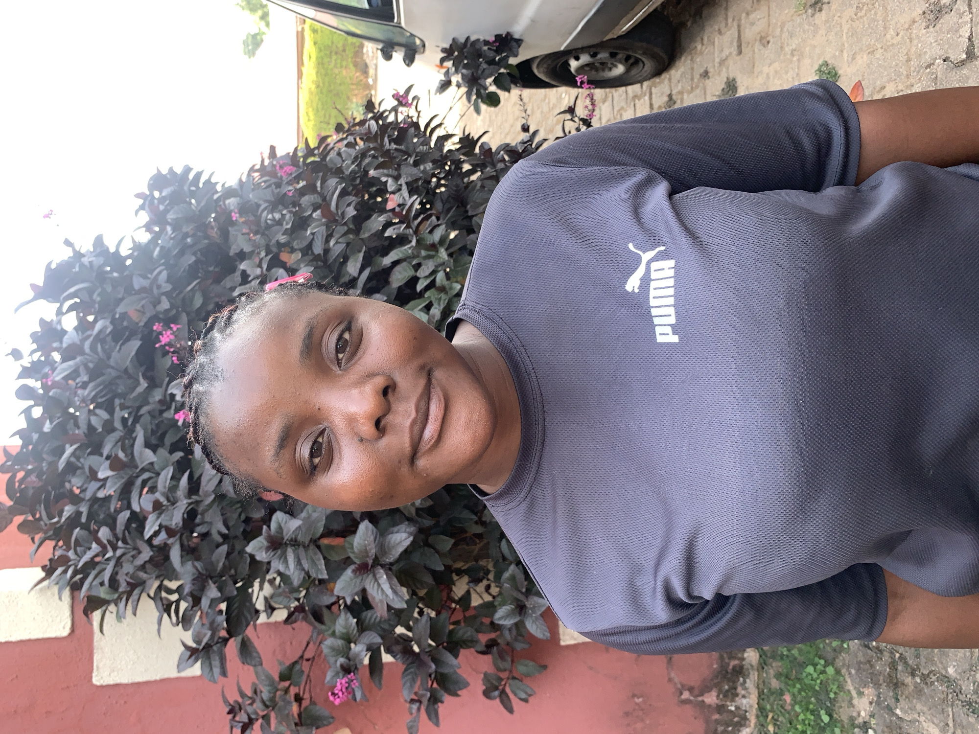 A portrait-style image, taken outdoors in Jos, Nigeria, features a young woman looking directly at the camera with a calm, friendly expression and a slight smile. She has dark skin and her hair is styled in small braids. She is wearing a dark grey, short-sleeved athletic t-shirt with the white "PUMA" logo and text prominently displayed on the chest.
The background includes lush, dark-leaved vegetation with small pink flowers, indicating a garden or natural setting. To the upper right, part of a white car, including a wheel, is visible behind the foliage. A reddish-brown wall with light-colored detailing runs along the left side of the frame. The ground appears to be paved or concrete. The scene is brightly lit by natural daylight, suggesting it was taken during the day.