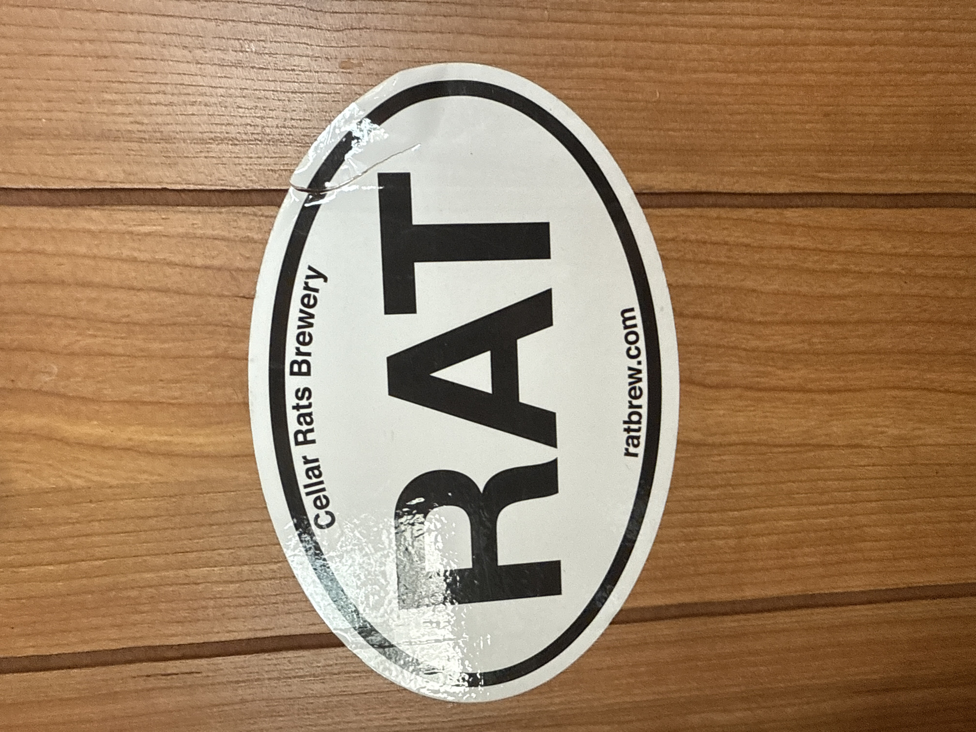 The image shows an oval-shaped sticker affixed to a wooden surface. The sticker has a black outline and the word "RAT" in large, bold, black letters in the center. To the left of "RAT" is the text "Cellar Rats Brewery," written vertically. To the right of "RAT" is the text "ratbrew.com," also written vertically. The sticker is peeling slightly at the top edge. The wooden surface has a horizontal grain pattern. The location, Davie, United States of America, is not discernable from the image.