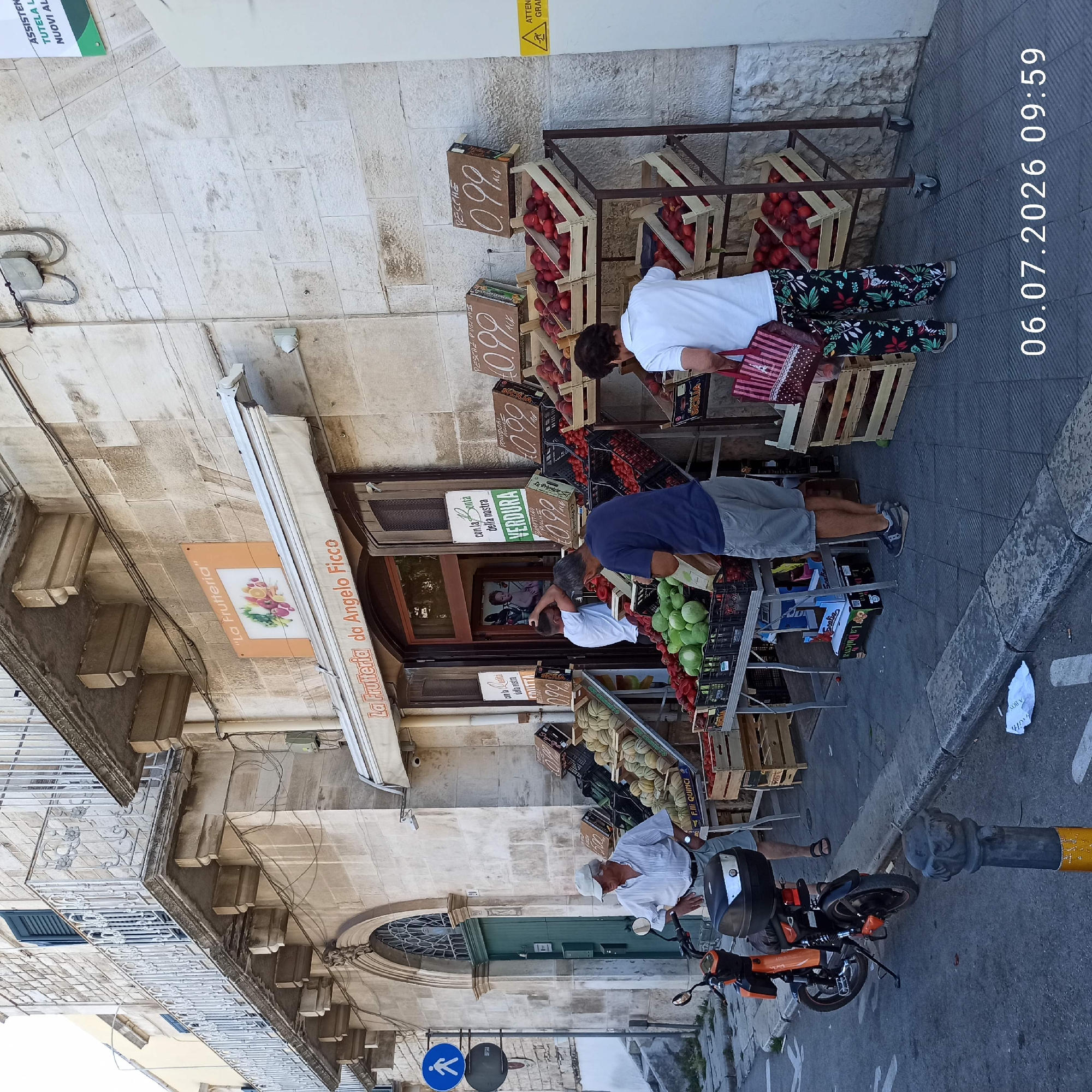 The image shows a fruit stand on a city street in Ruvo di Puglia, Italy. The fruit stand is set up in front of a building with stone facade and a balcony. There are several people present. An older man wearing a white striped shirt, light-colored shorts, and a white cap stands near a scooter. Another man, wearing a blue t-shirt and gray shorts, is examining produce at the stand. A person, whose face is not visible, wearing a white t-shirt and floral patterned pants, is shopping at the stand and holding a bag. The fruit stand displays a variety of fruits and vegetables, including melons, tomatoes, peaches, and what appear to be gourds or large green pears. Wooden crates are used to hold the produce, and price signs are visible. An orange scooter is parked on the sidewalk. The environment appears to be a sunny day. The street is paved, and there is a curb and sidewalk. A blue pedestrian crossing sign is visible on a pole. The building above the fruit stand has an awning and a sign that reads "La Frutteria da Angelo Ficco".