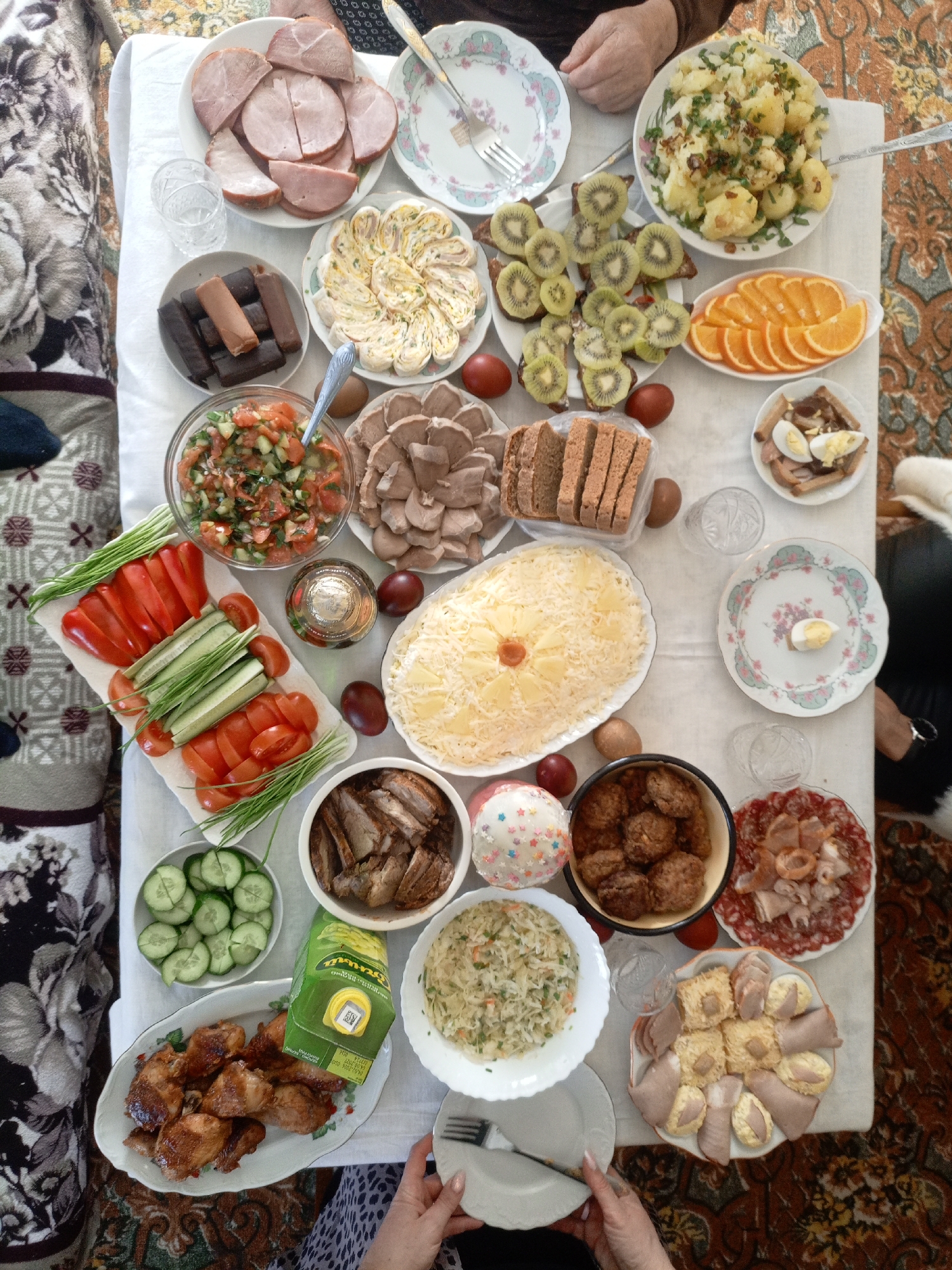 The image shows a table laden with food, seemingly in a home setting within the Лунненскі сельскі Савет region of Belarus. The table is covered with a white tablecloth and displays a variety of dishes including sliced ham, what appears to be a vegetable salad, sliced cucumbers and tomatoes, cooked meat, sliced bread, kiwi slices on bread, orange slices, a rice dish garnished with pineapple, meatballs, and a dessert item. A few people are visible around the table, with hands reaching out to take food. Various plates and forks are scattered around, suggesting a gathering of people sharing a meal. Some drinks are also on the table. The setting is indoors, likely a dining room, as evidenced by the tablecloth and assorted food items. Furniture is visible in the background, including a sofa with a patterned cover. A patterned rug is underneath the table. Overall, it appears to be a casual gathering with an abundance of food and drink.