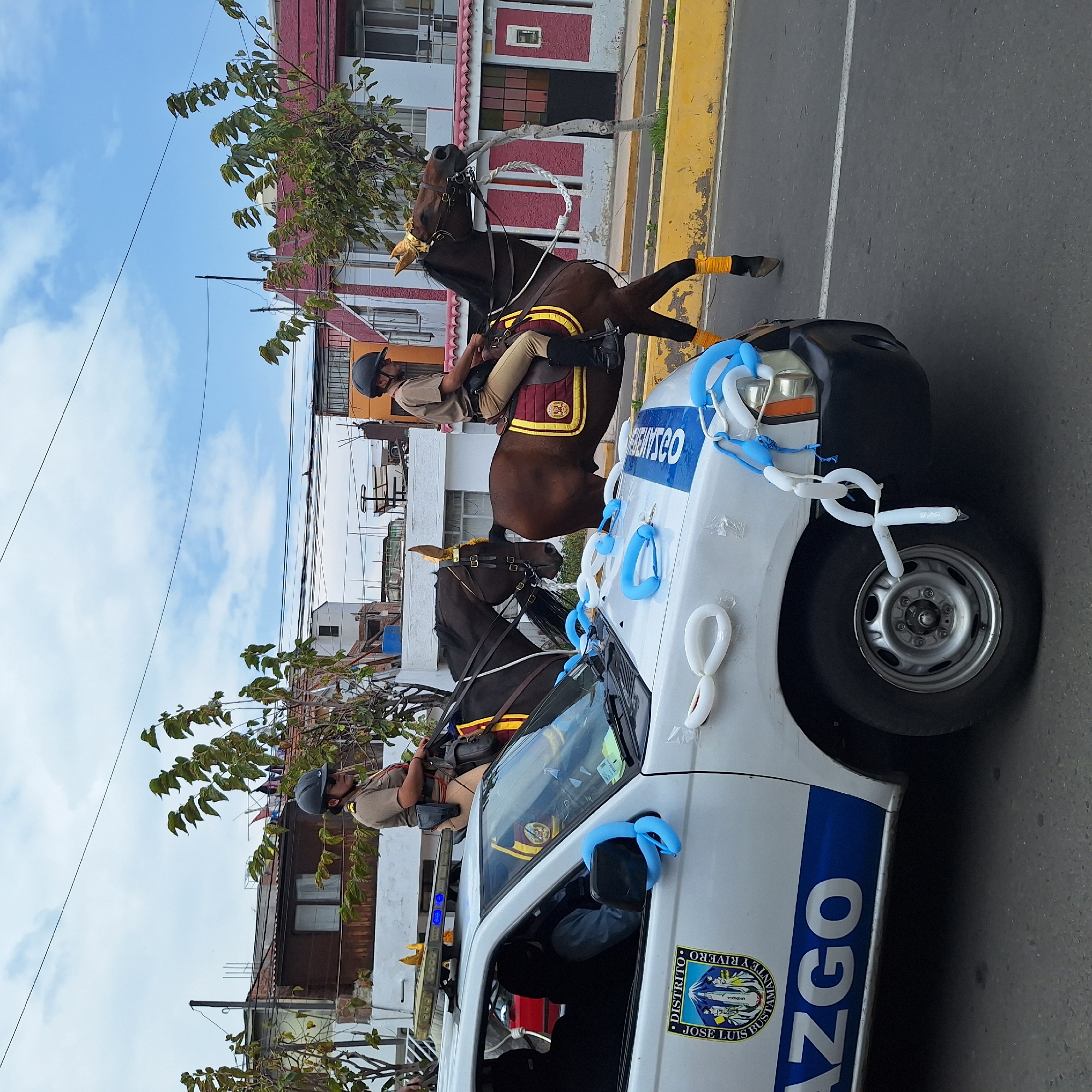 The image depicts two mounted police officers on horseback next to a white police vehicle decorated with blue and white balloons. The vehicle is marked with "AZCO" and "DISTRITO JOSE LUIS BUSTAMANTE Y RIVERO", confirming the location as José Luis Bustamante y Rivero in Peru. The officers wear helmets and uniforms, and the horses have decorative yellow accents. The scene appears to be part of a parade or public event, situated on a street with buildings and trees in the background. The sky is visible with scattered clouds.