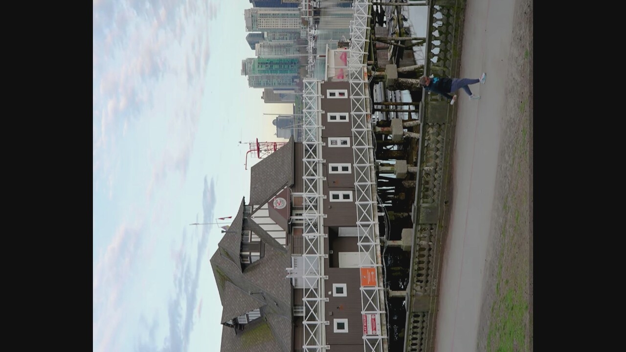 A person is jogging on a paved path in the foreground. Behind them, a brown building with white railings is situated on stilts over the water, with a cityscape visible in the background. The building appears to be a boathouse or similar structure, with a white fence running along its length. The water is calm, reflecting the sky and the surrounding buildings. The sky is overcast with soft clouds. In the distance, a modern skyline with numerous skyscrapers dominates the horizon. The overall scene suggests an urban waterfront environment. The boathouse is located in Coal Harbour, Vancouver, Canada.