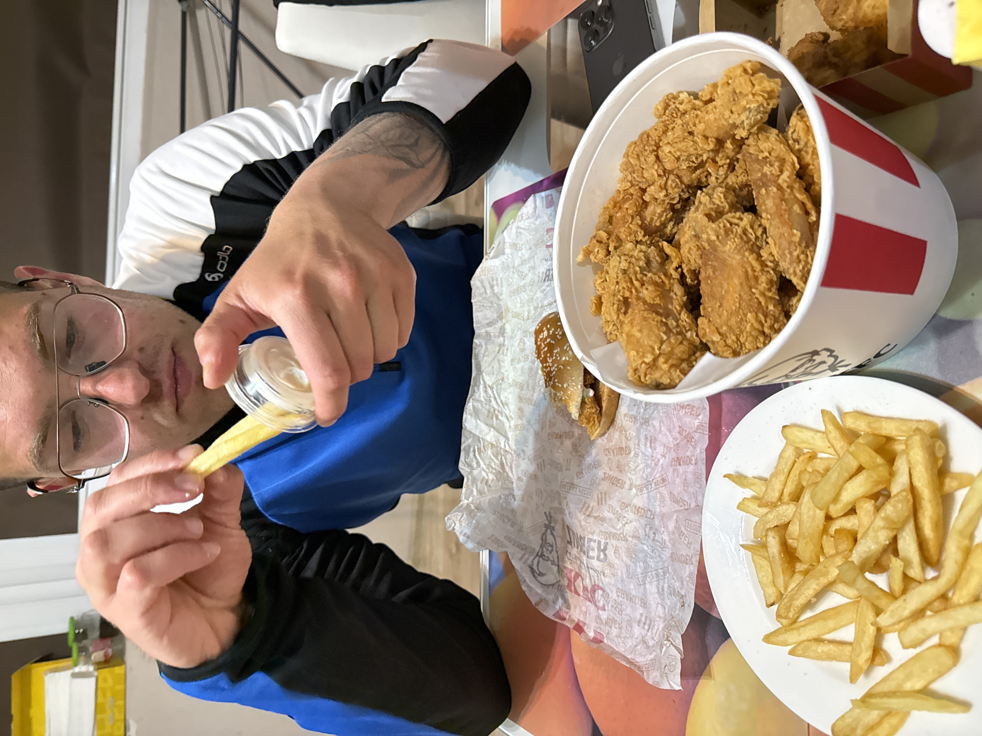 A man wearing glasses and a blue and black jacket is shown in an indoor setting, pouring sauce on a french fry. A bucket of fried chicken, a plate of french fries, and a sandwich in KFC paper wrapping are on the table. A black smartphone is present. The location is unknown.