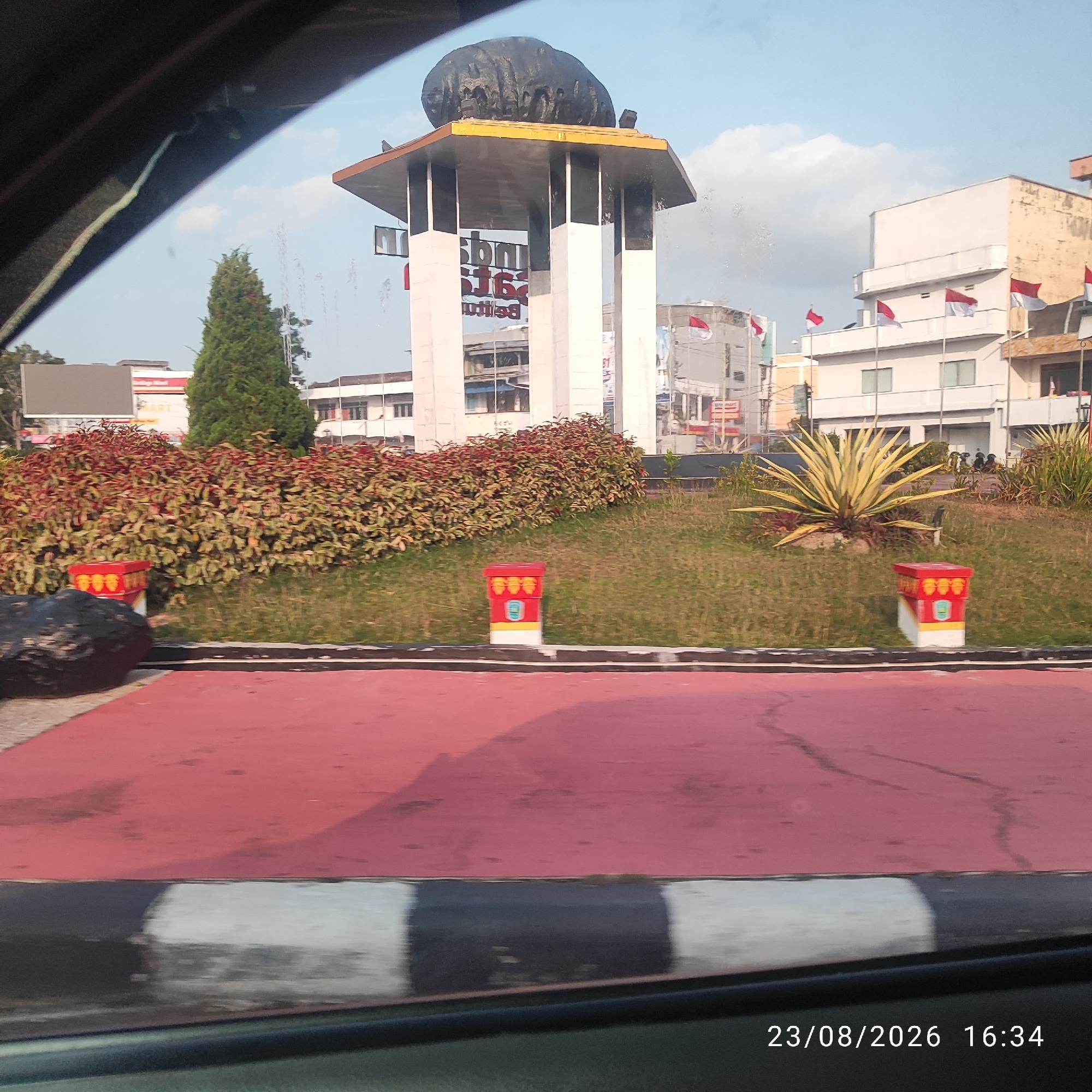 The image captures a street view in Tanjung Pandan, Indonesia, likely a prominent landmark based on the monument. In the foreground, a red road surface with black and white striped borders is visible, suggesting it's a pedestrian area or a median. To the left, a dark, irregular-shaped rock sits on the red surface. In the mid-ground, a meticulously maintained garden features a large, spiky, yellow-green plant and a dense hedge with red and green foliage. Two decorative red and white cylindrical structures with yellow accents are placed within the garden.
Dominating the background is a large, multi-tiered monument with white pillars supporting a gold-colored roof. Atop the monument rests a large, dark, boulder-like sculpture. White buildings with various architectural styles are visible behind the monument and surrounding greenery, some adorned with Indonesian flags. A tall, conical evergreen tree stands to the left of the monument. The sky is a clear blue with a few scattered white clouds. The overall scene suggests a public space, possibly a roundabout or plaza, in a well-kept urban environment. The timestamp in the lower right corner reads "23/08/2026 16:34".