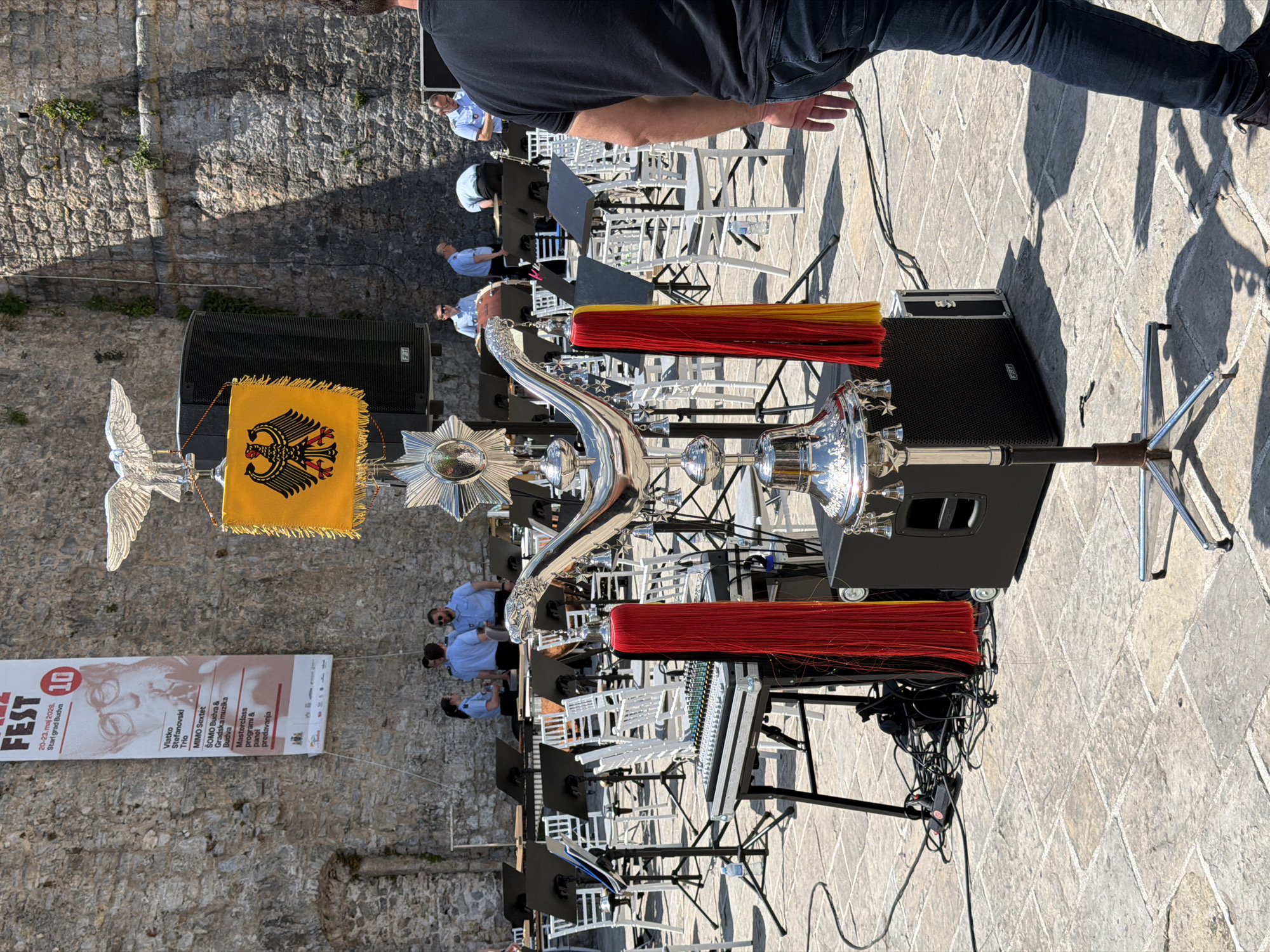 The image captures an outdoor scene in Budva, Montenegro, specifically within the historic "Stari Grad" or Old Town. A music band is setting up or has just finished performing, with instruments and equipment such as speakers, chairs, and music stands arranged on a stone-paved surface. A banner with the text "FEST 20-23. maj 2026. Stari grad Budva" and "Vlatko Stefanovski Trio" hangs on the weathered stone wall in the background, hinting at a cultural festival or event. Several men are seen wearing light blue shirts and dark pants, appearing to be members of the band or crew. The presence of a flag with an eagle emblem and ornate silver musical instruments suggests the band has a formal or ceremonial role. The sky is bright, indicating a sunny day. A mixing console is visible with its wiring and cables in disarray. A person wearing jeans is standing nearby observing the scene.