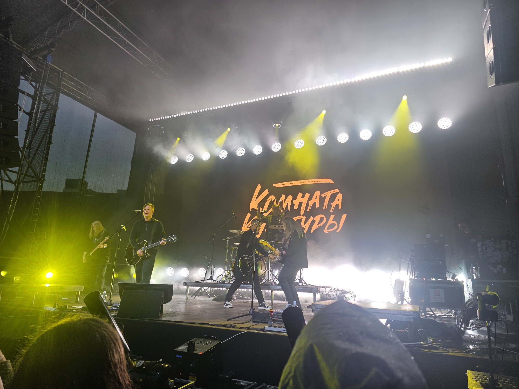 The image shows a rock concert performance on an outdoor stage in Kazan, Russia, under a bright, yellow-lit sky. The stage is equipped with professional lighting, speakers, and musical instruments. In the background, a large screen displays the band's name in orange Cyrillic script, which reads "КОМНАТА КУЛЬТУРЫ" (Komnata Kultury). The band members are actively performing, with two guitarists and a drummer visible on stage. One guitarist is in the foreground on the left, playing a black electric guitar, while another guitarist is positioned more centrally, also playing an electric guitar. A drummer is seated behind his drum kit in the background. The audience is partially visible in the foreground, with the backs of their heads and shoulders showing, suggesting they are standing and watching the performance. The atmosphere is energetic, with stage smoke and bright lights creating a dramatic effect.