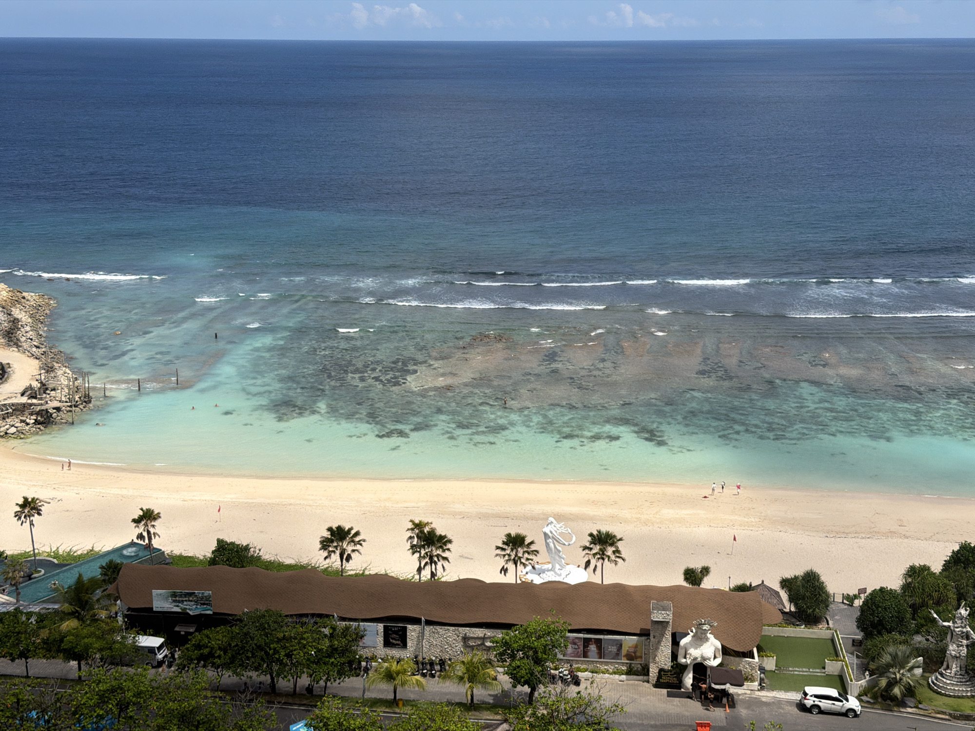 The image depicts a high-angle view of Melasti Beach in Ungasan, Bali, Indonesia. The scene showcases a clear, sunny day with a blue sky and a portion of the ocean that transitions from a darker blue to turquoise near the shore. Waves are visible as they break near the beach.
A sandy beach stretches across the mid-ground. Several palm trees are scattered along the coastline, near a building with a brown, undulating roof. White statues are positioned on the beach and at the building's entrance.
Near the building, there are trees and shrubs, along with a paved road where a white car is parked. A few people are seen walking and swimming near the beach.